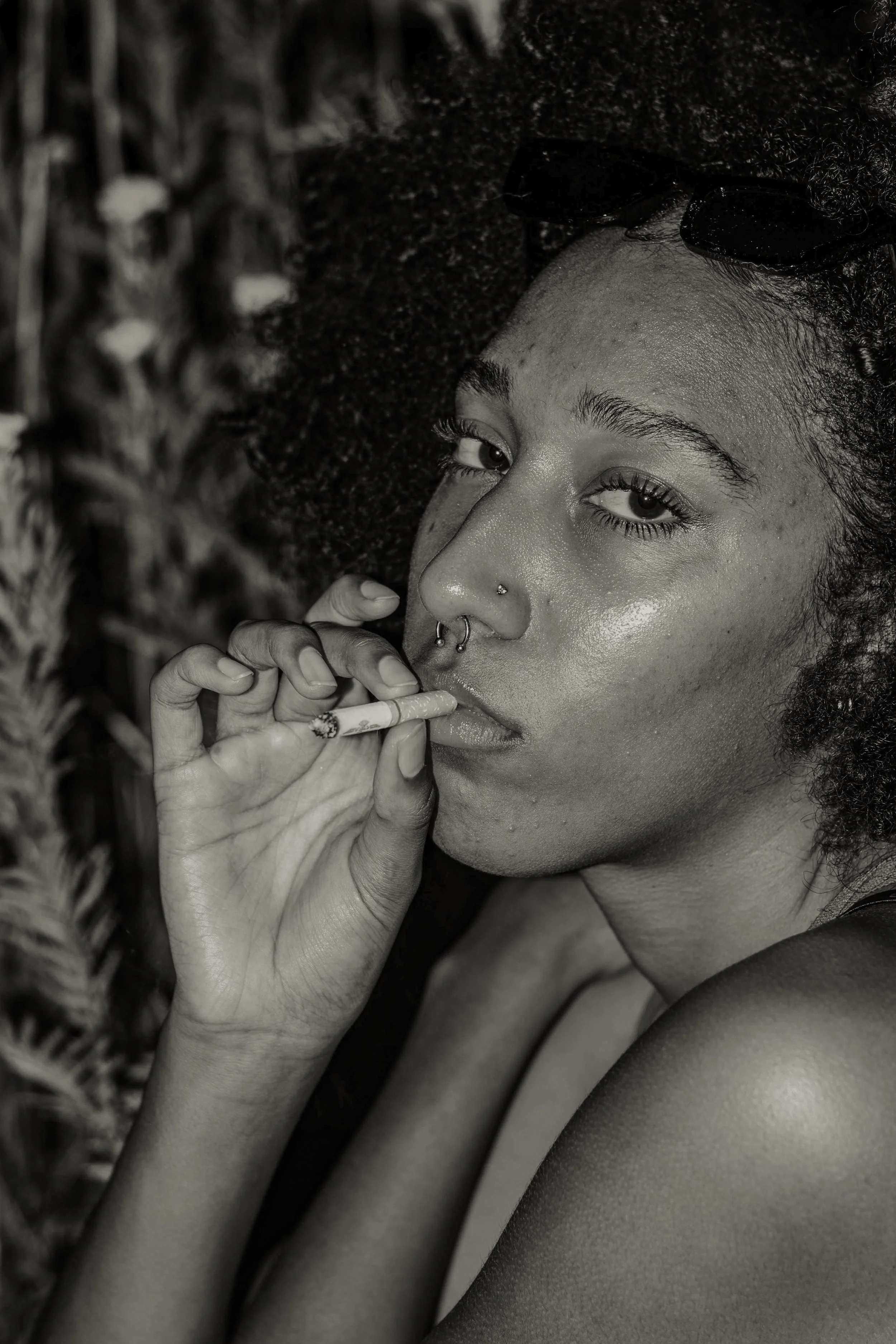 A young woman with curly hair, sunglasses on her head, and nose piercings holds a cigarette near her lips while looking into the distance.