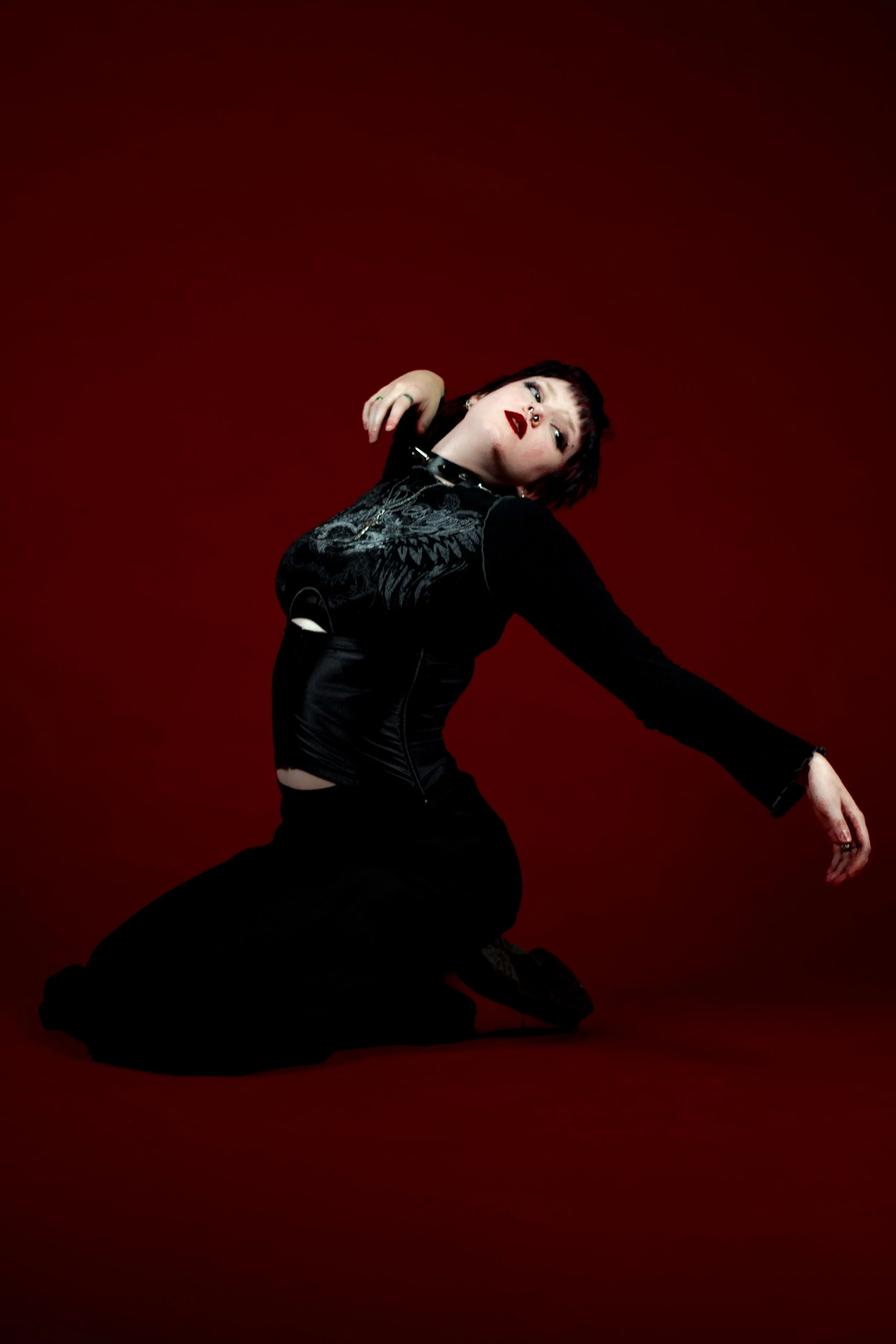 Woman with short dark hair and dark makeup wearing a black patterned jacket and black pants, posing against a maroon background.