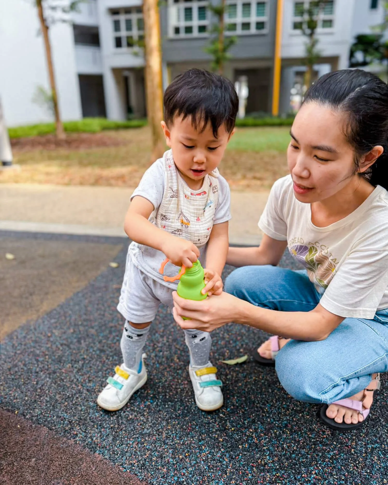 A woman helps a young boy open a green water bottle at a playground, both are kneeling on colorful rubber flooring outdoors.