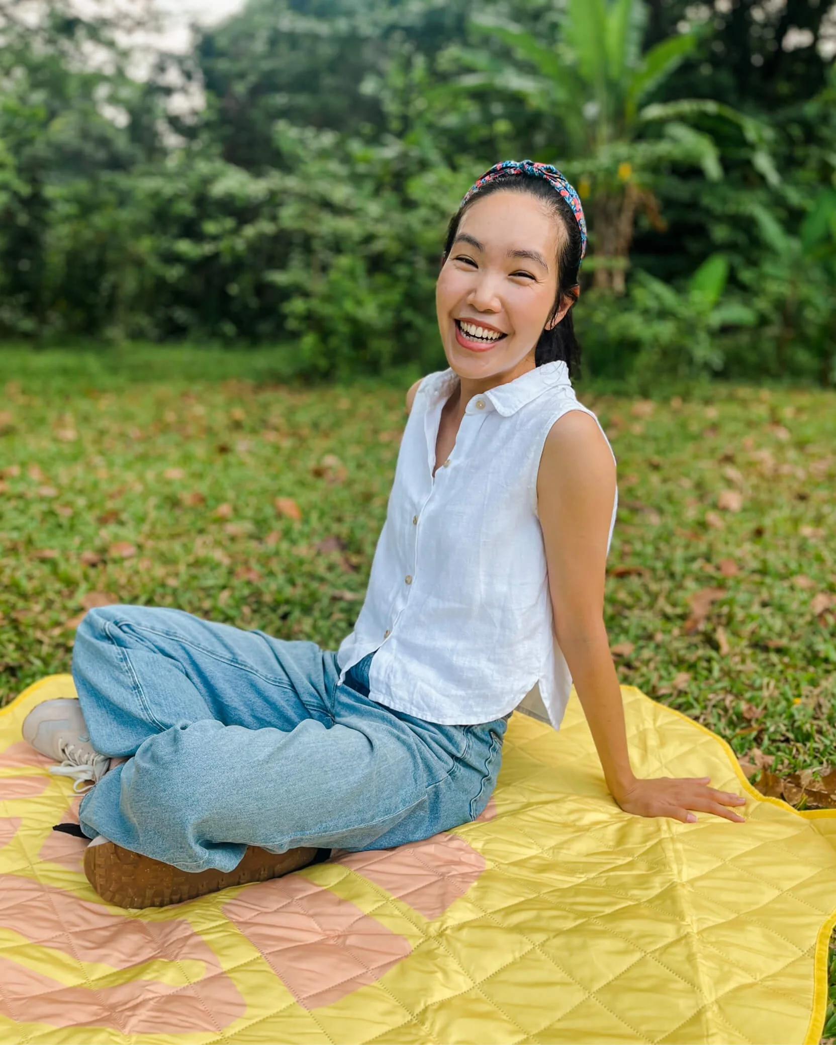 Woman sitting on a yellow picnic blanket, smiling, outdoors on grass with trees and greenery in the background.