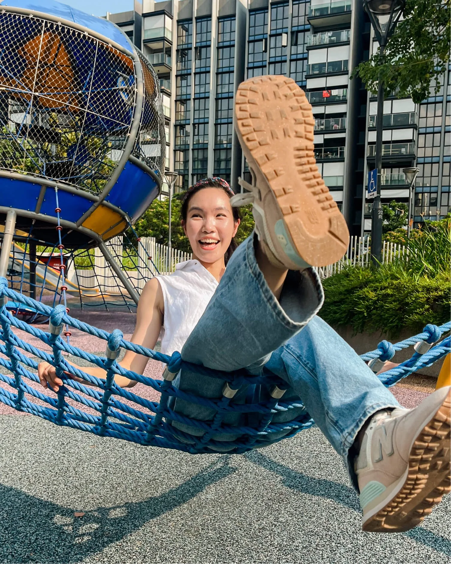 A smiling person reclines on a blue rope hammock at a playground, with buildings and play structures in the background.