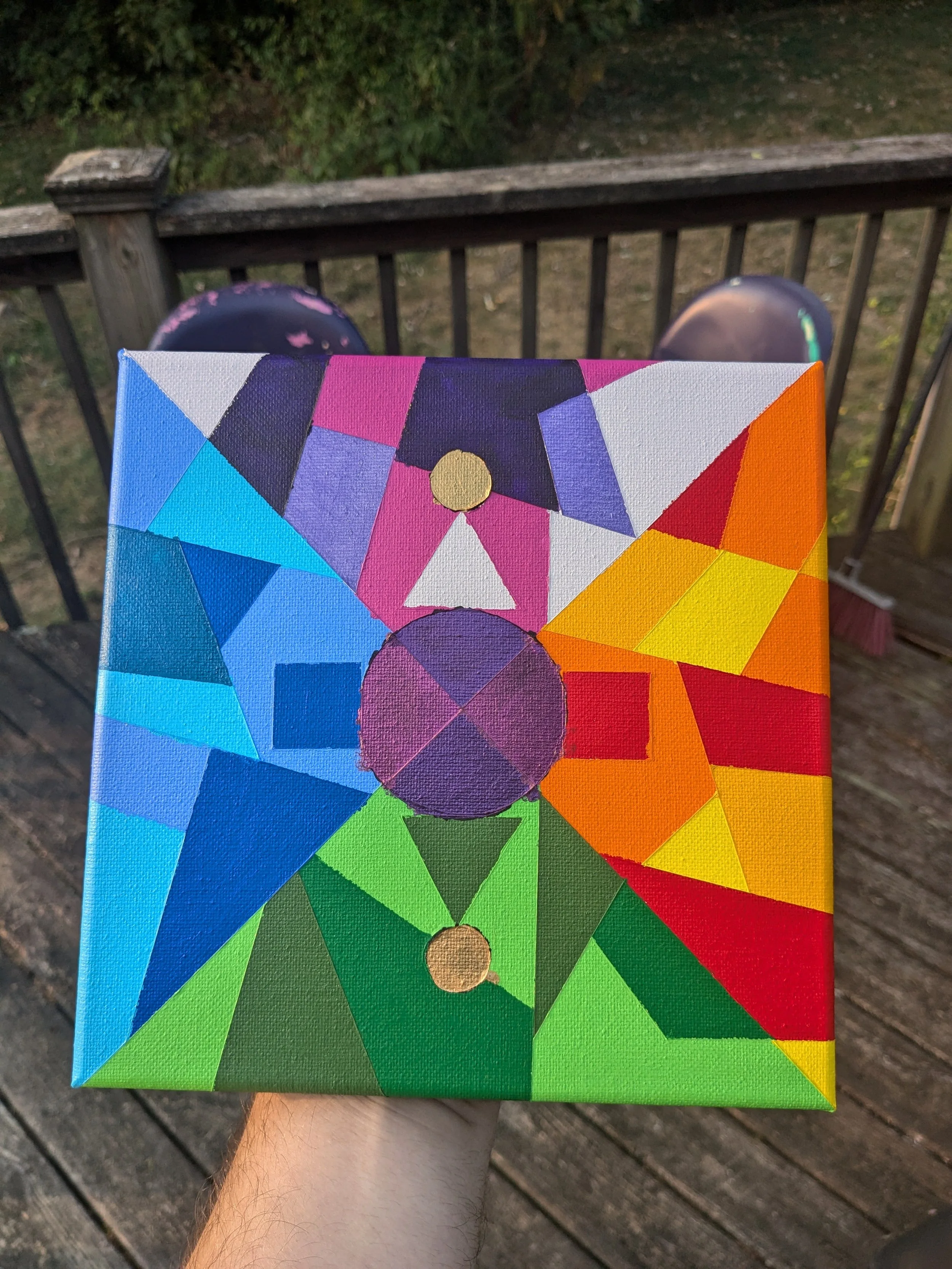 A hand holding a colorful, geometric abstract painting outdoors on a wooden deck, with a background of foliage and a railing.