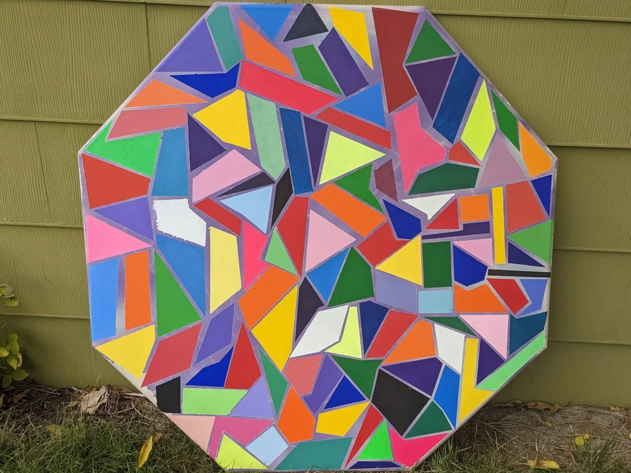 A colorful mosaic art piece shaped like the state of Alabama, composed of various brightly colored geometric triangles and shapes, mounted outdoors against a green wooden fence.
