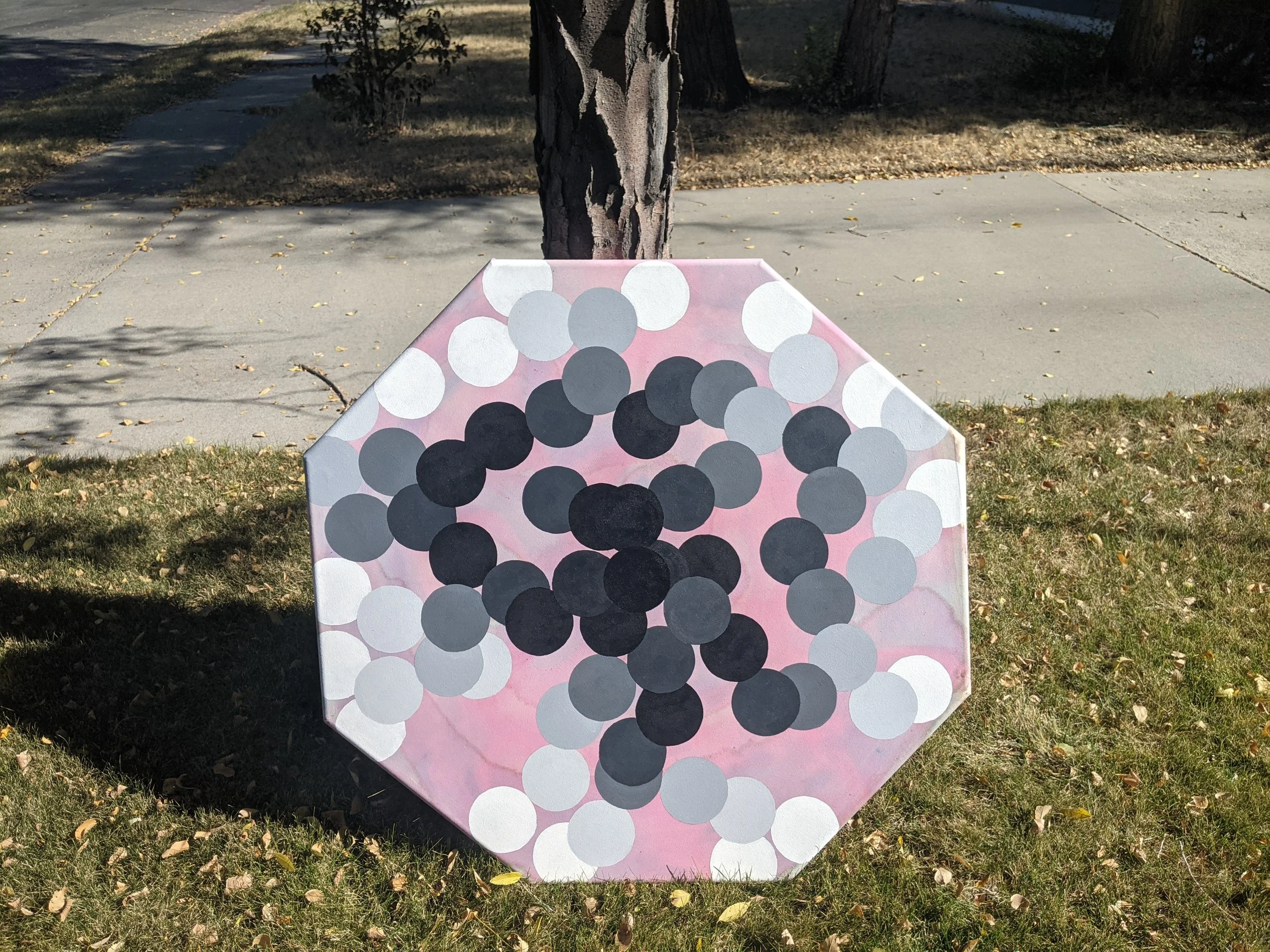 A painted object resembling a cluster of black and gray circles on a pink background, placed on grass beside a sidewalk and a tree in a park or yard.