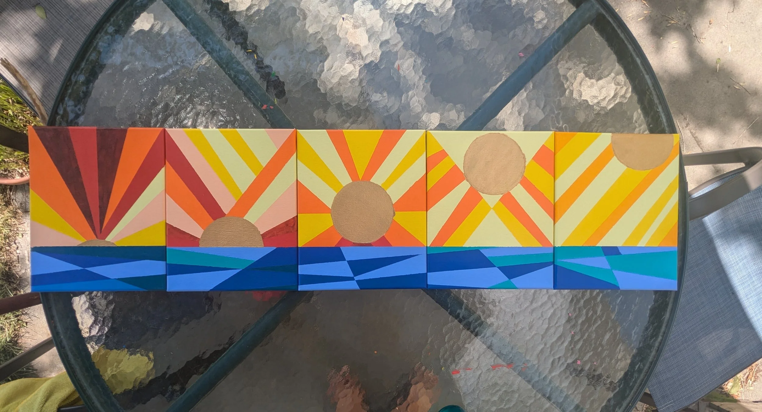 Colorful geometric abstract art consisting of six square panels, each with sun, rays, and water designs in vibrant colors including orange, yellow, pink, blue, and brown, arranged in a horizontal row on a glass table.