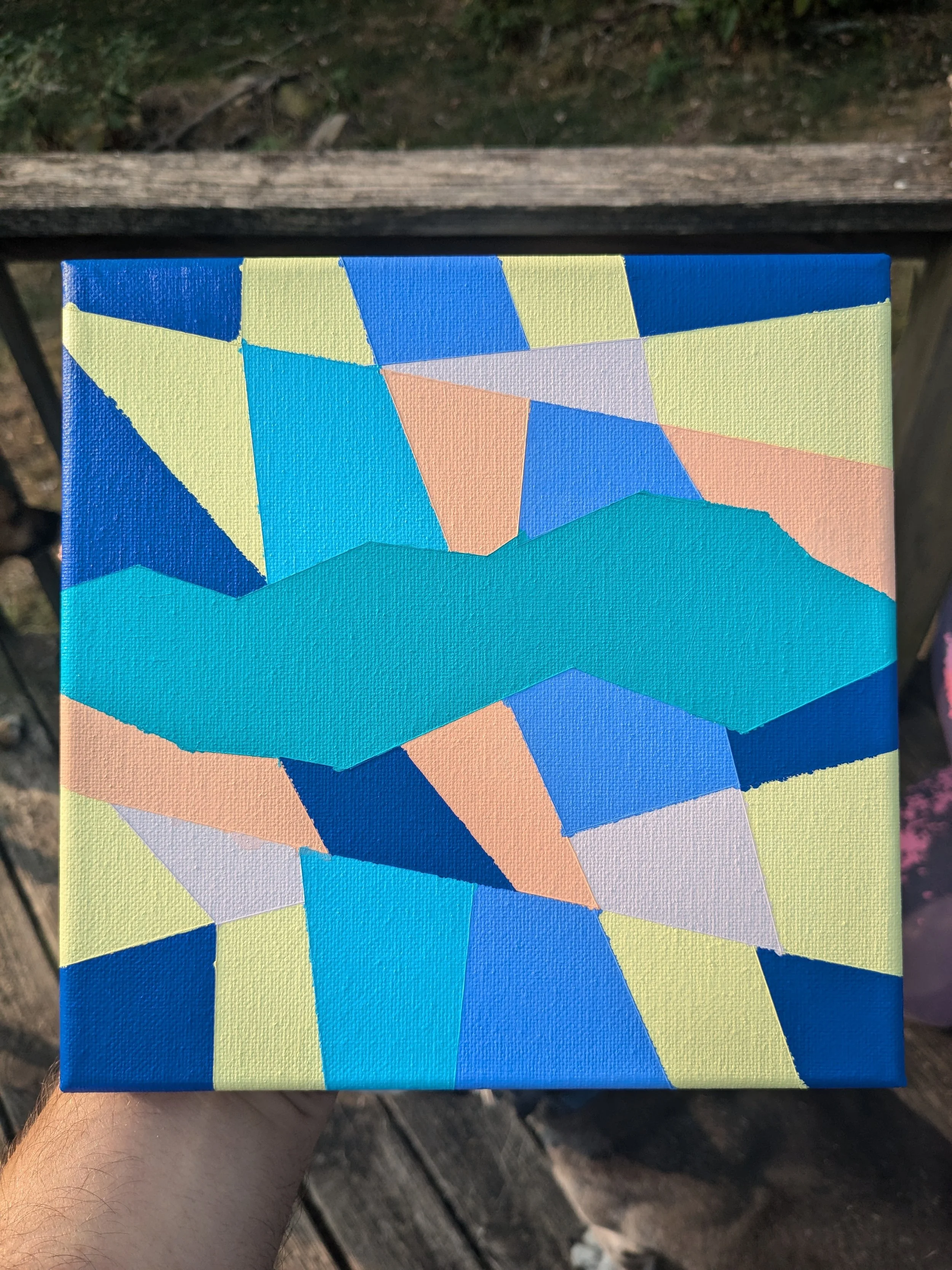 Close-up of a colorful abstract geometric painting on a small canvas held in a person's hand outdoors, with a wooden surface and green foliage in the background.