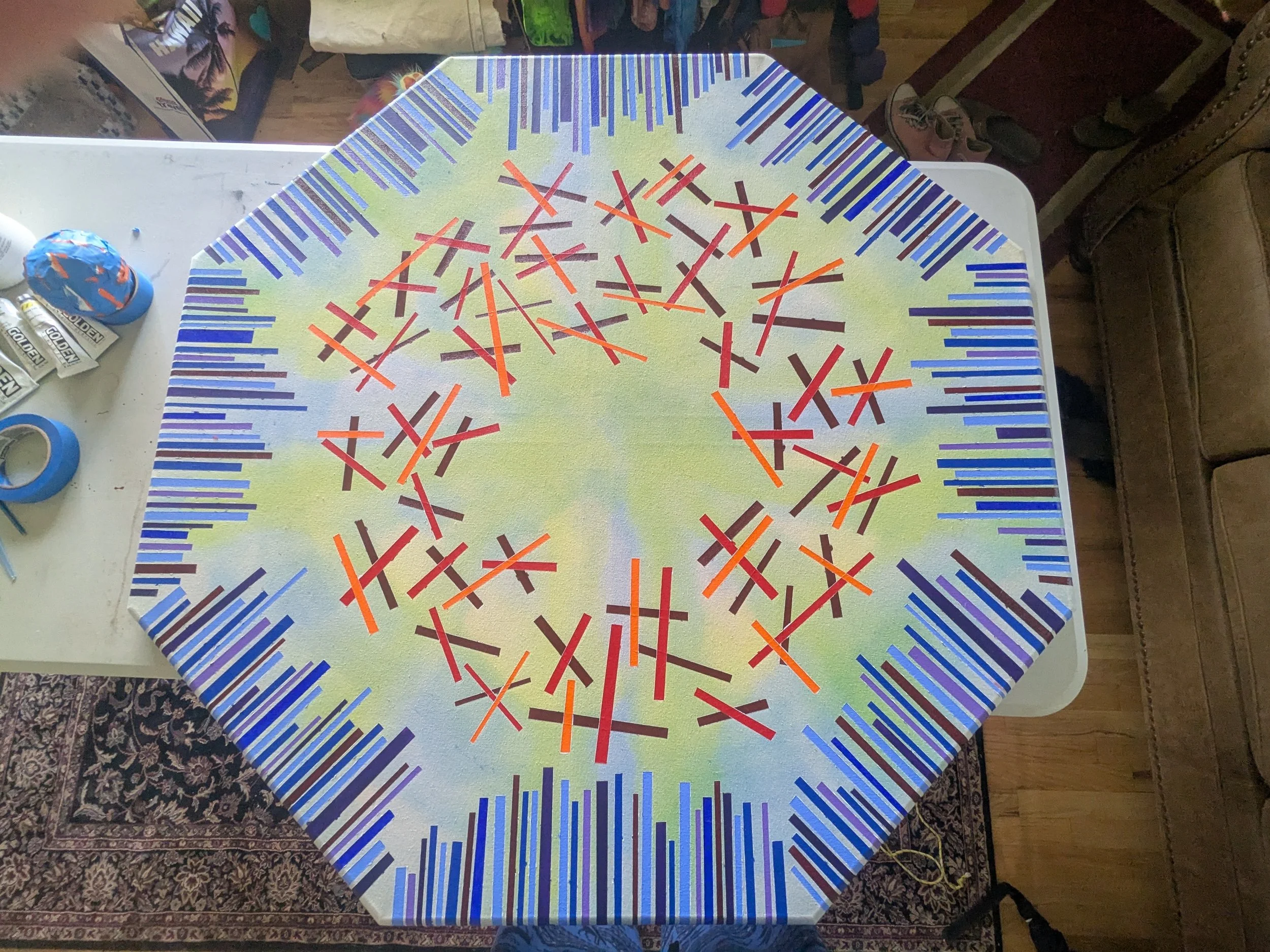 A table with a colorful Zigzag pattern tablecloth featuring red, orange, purple, and blue lines. In the background, there are craft supplies including tape, glue, a paper roll, and a paint bottle, with a patterned rug underneath the table.
