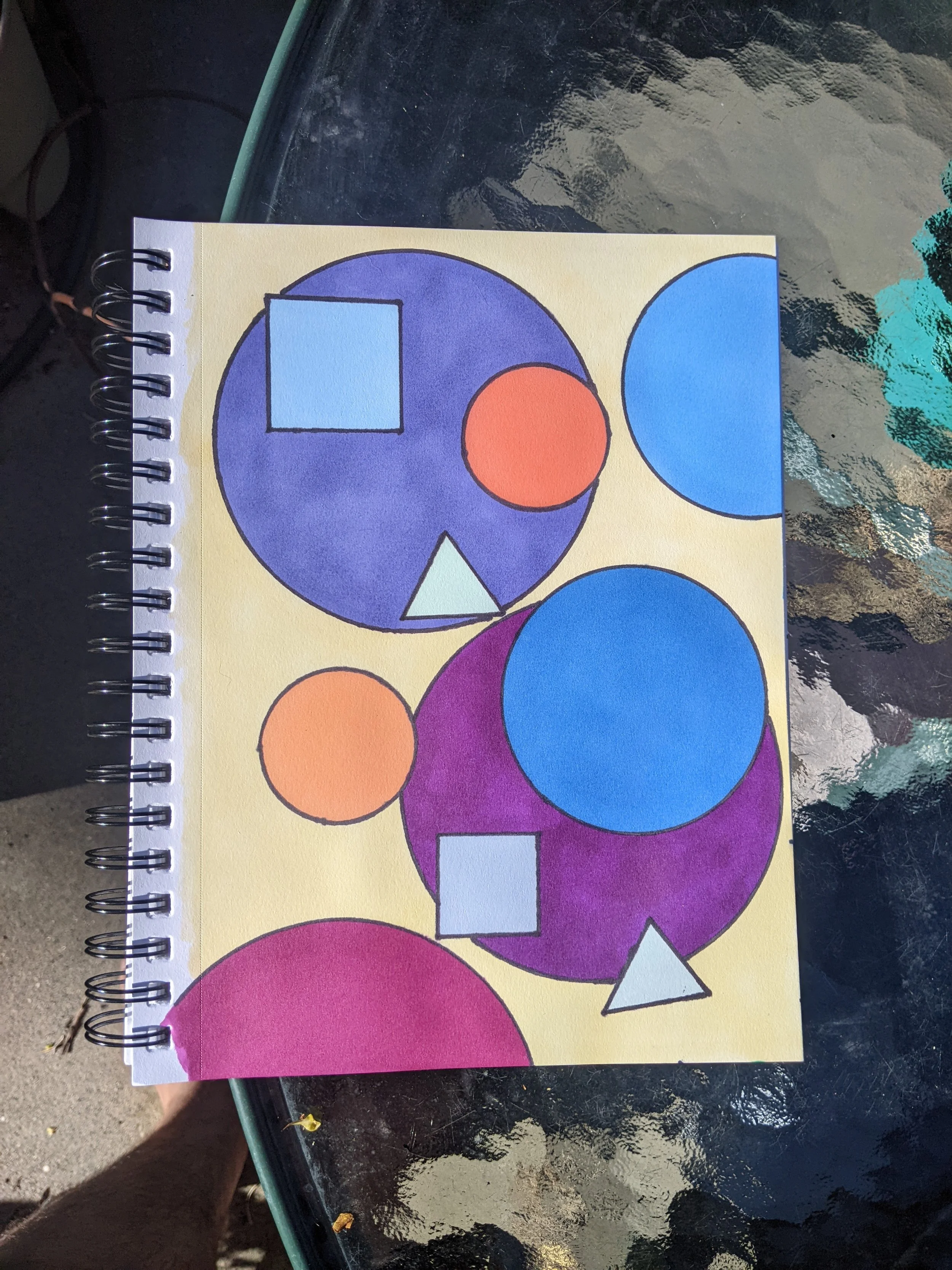 A colorful abstract design with various shapes including circles, squares, and triangles outlined in black on paper. The background of the paper is yellow.