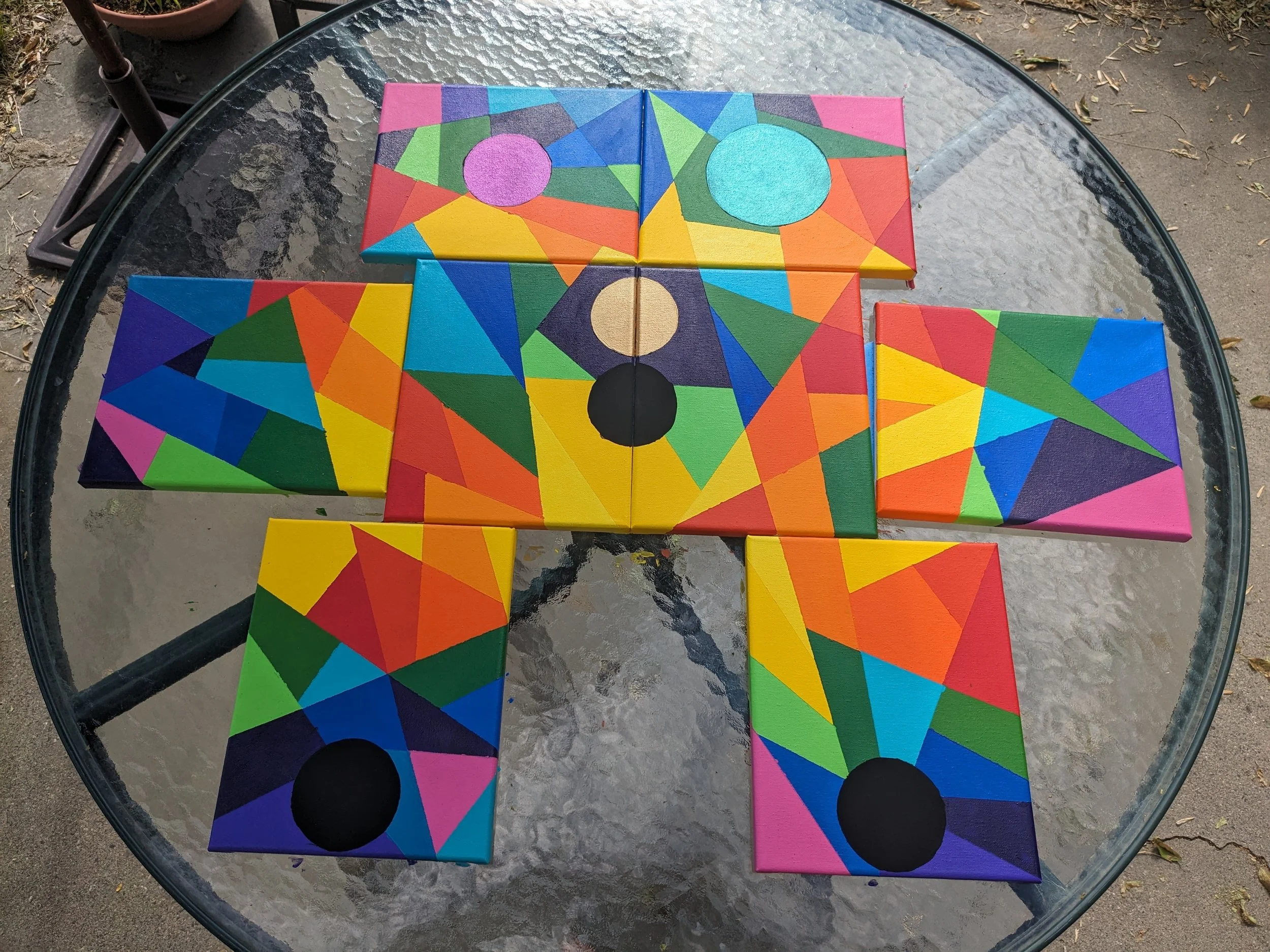 Colorful geometric abstract art painted on multiple canvas panels, arranged on a glass outdoor table, featuring various shapes and vibrant colors.