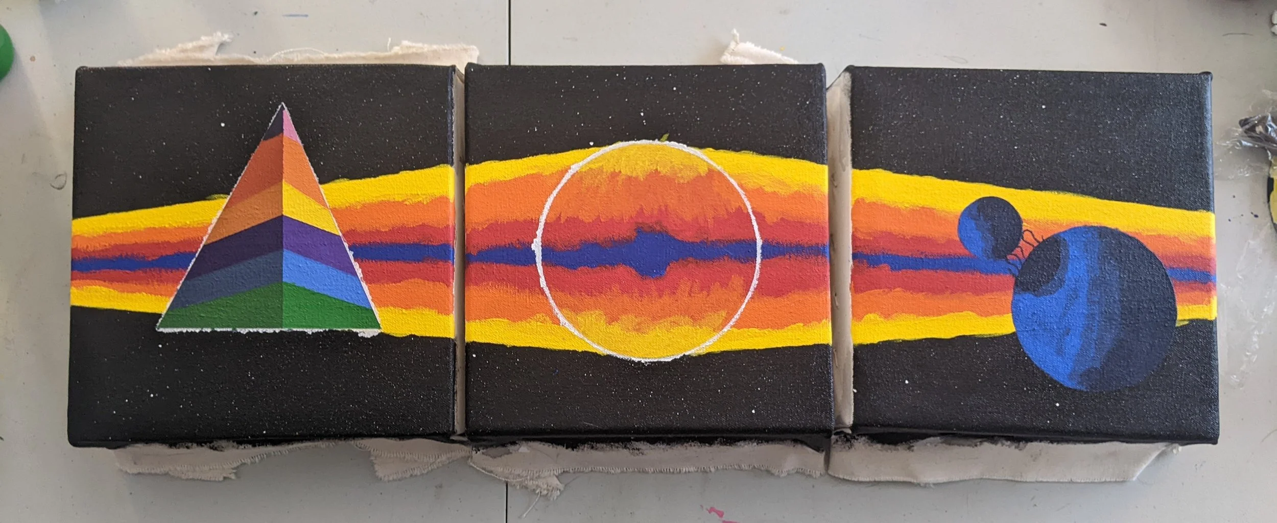 Triptych painting on three canvas panels features a black background with horizontal streaks of yellow, red, orange, and blue across each panel. On the left panel, there is a geometric, pyramid-shaped object with rainbow stripes. The middle panel has