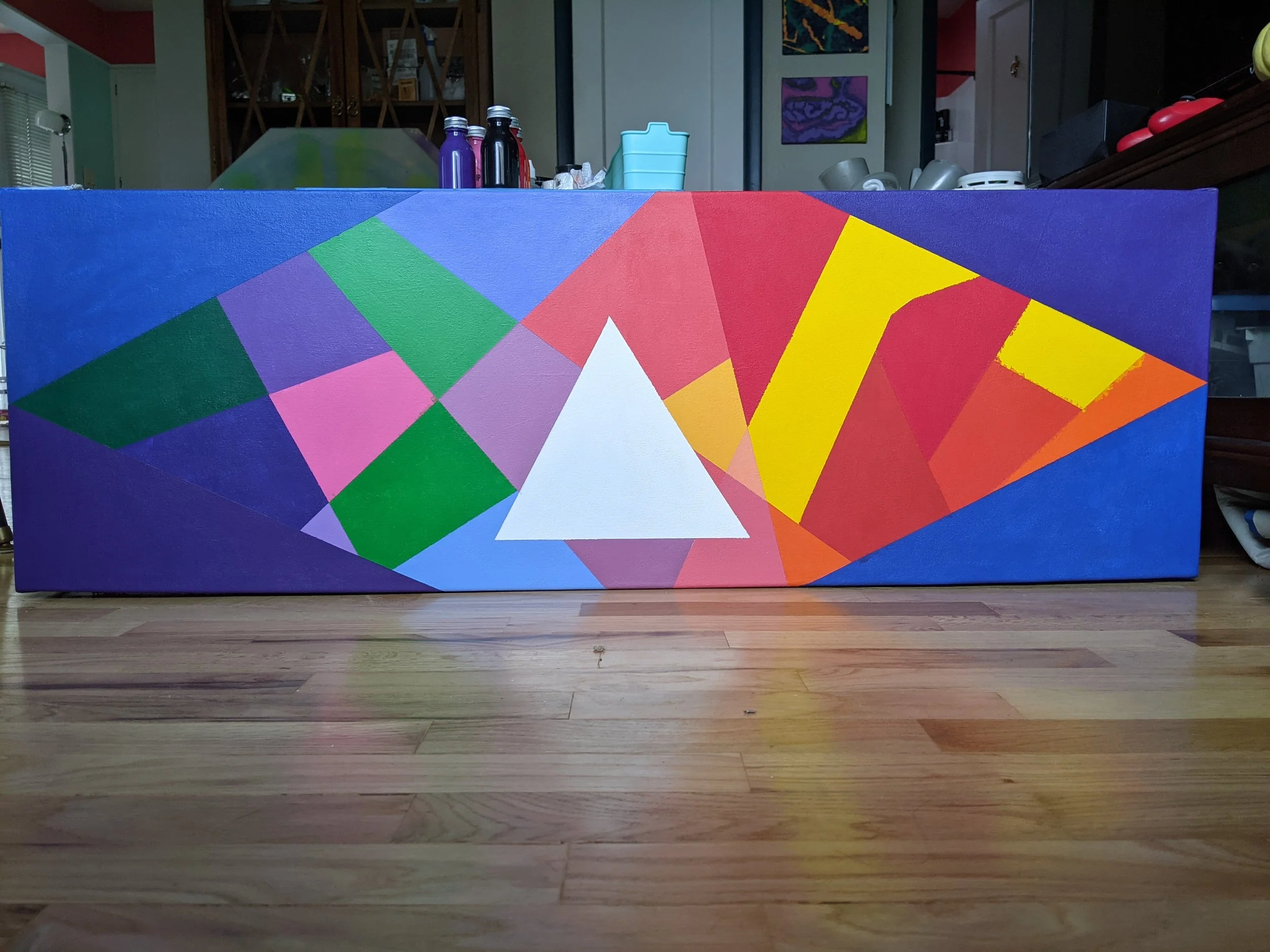 Colorful abstract geometric painting with various shapes and colors including blue, green, pink, red, yellow, purple, and white, placed on a wooden floor.
