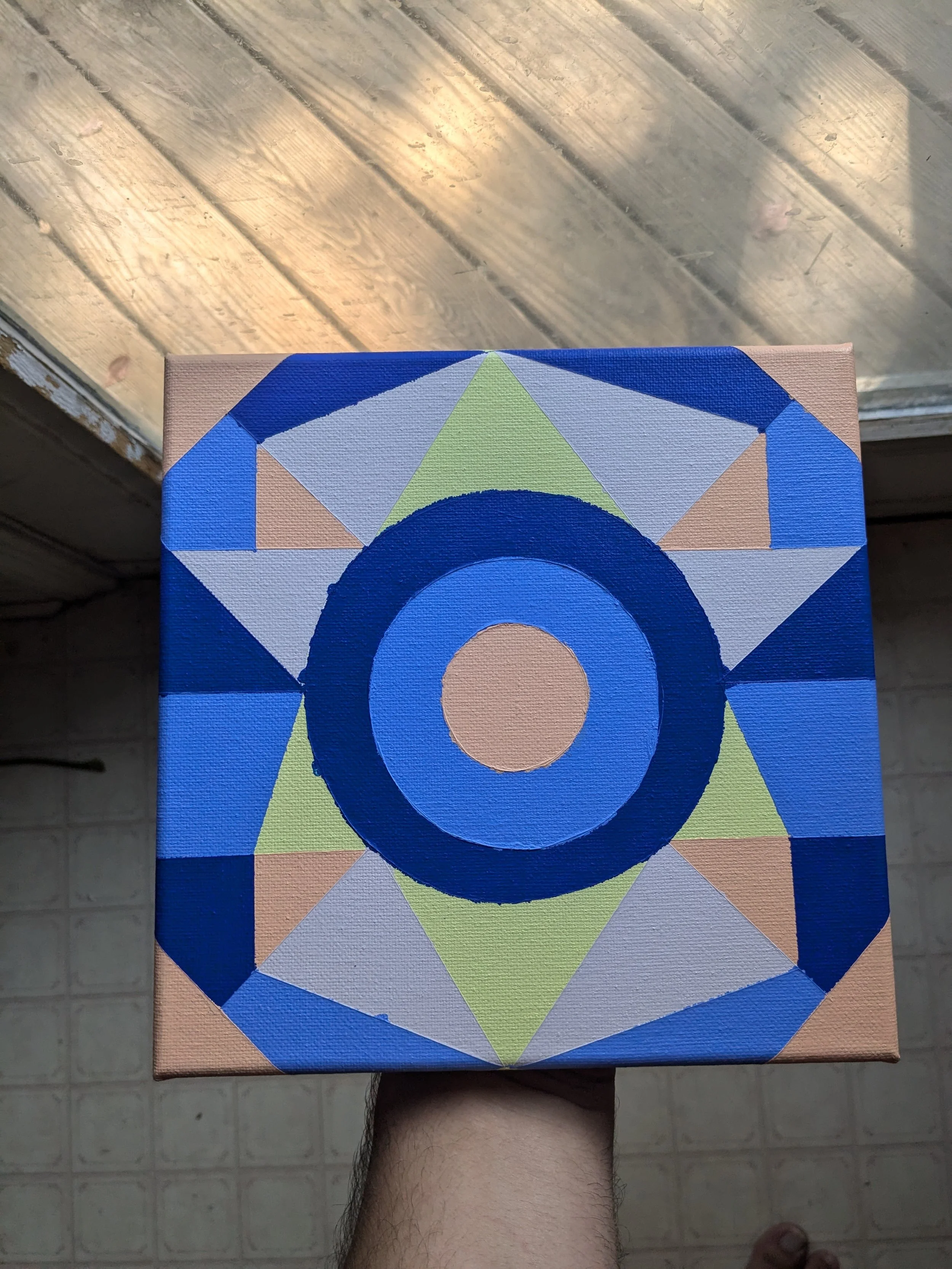 A hand holding a canvas with a geometric abstract painting featuring circles and triangles in blue, green, beige, and peach colors. The background is a wooden floor.