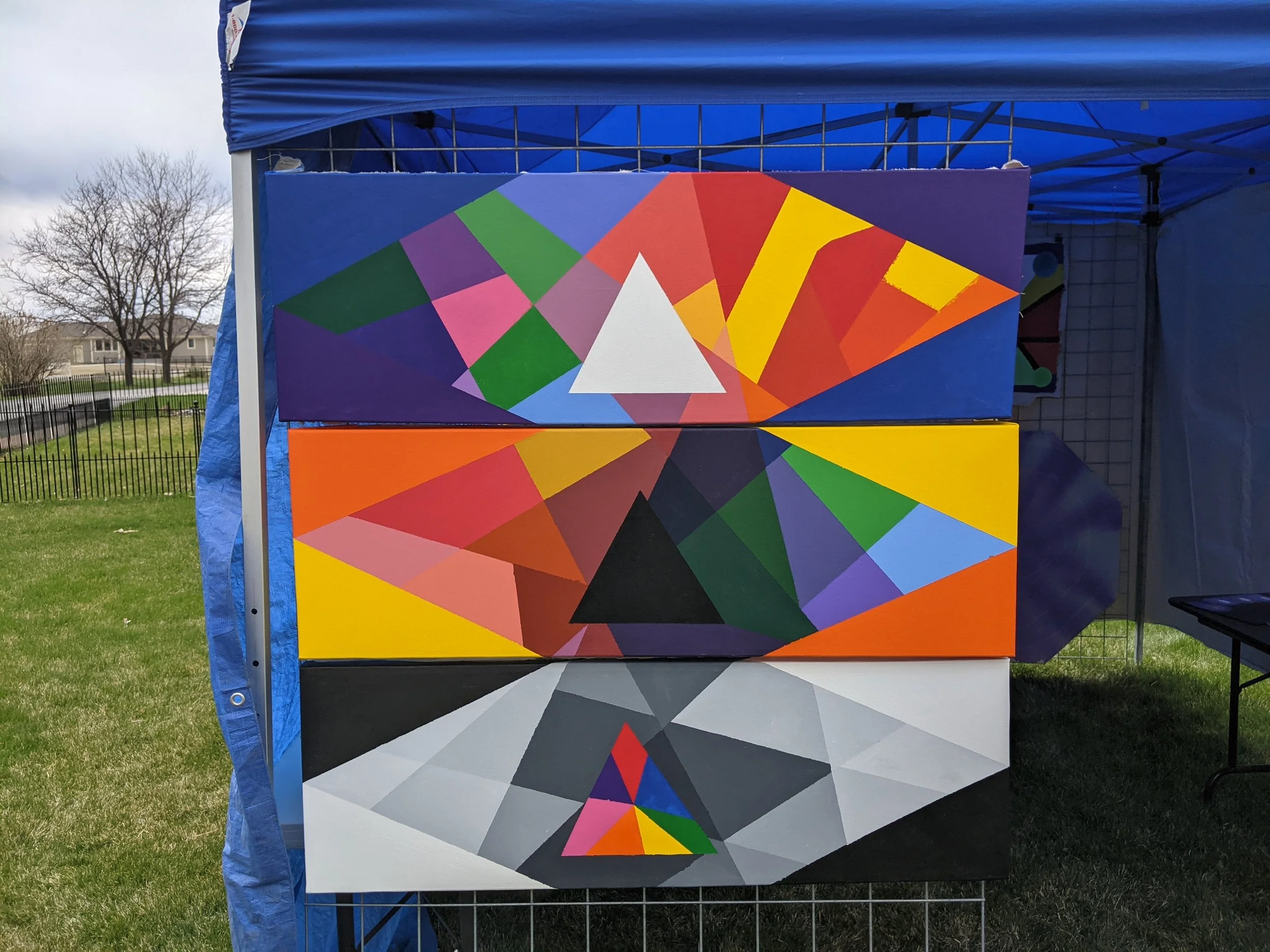 Three colorful geometric paintings on display under a blue tent at an outdoor event with grass and trees in the background.