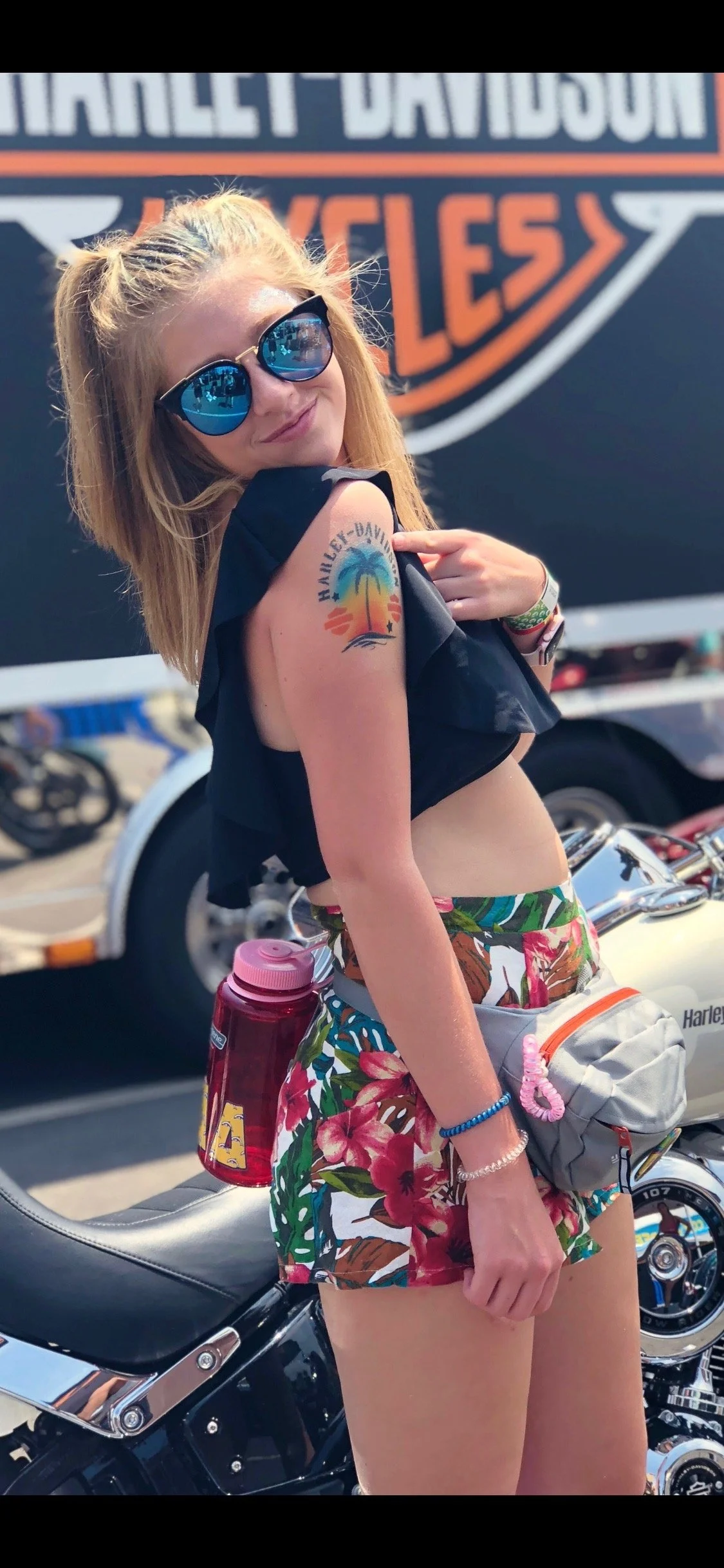 A young woman with blonde hair and sunglasses showing a Harley-Davidson temporary tattoo on her upper arm at a motorcycle event.