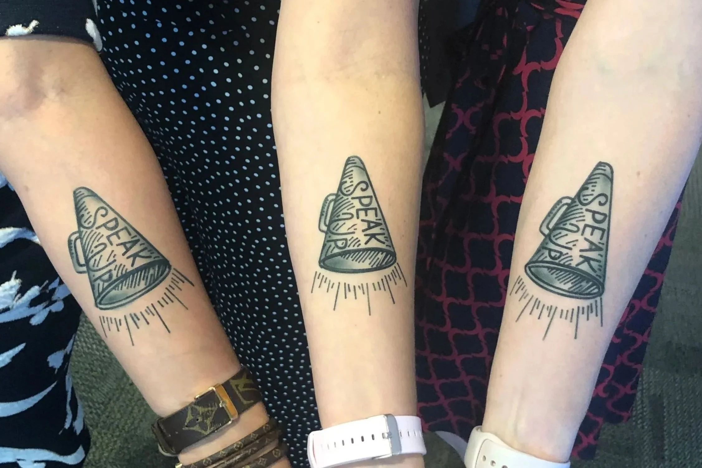 Four people show their forearms with matching temporary tattoo designs labeled 'SPEAK UP.' They are wearing different clothing, including polka dots and patterned dresses, and various wrist accessories.