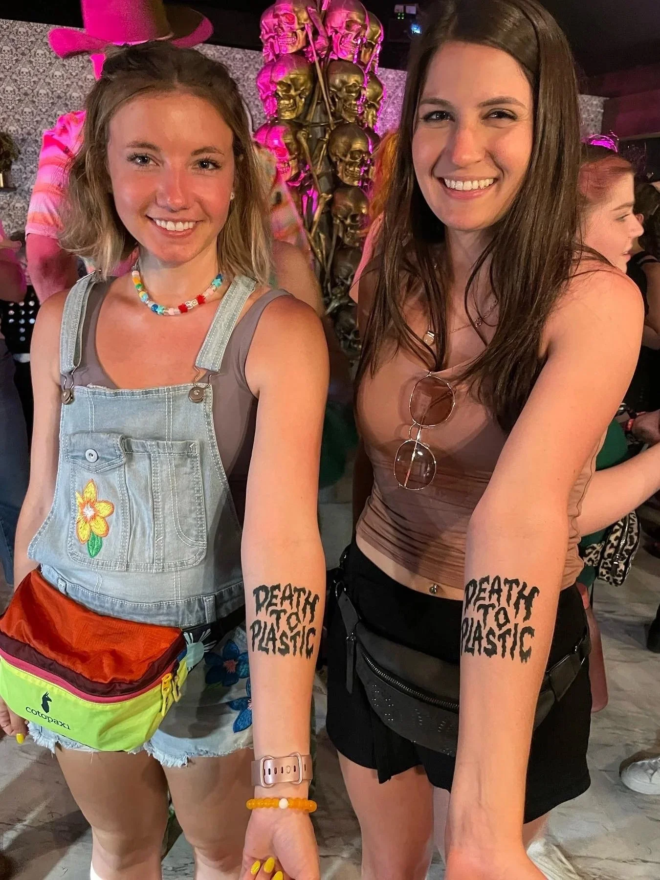 Two women standing side by side smiling, with a background of skull decorations. They have temporary tattoos on their arms that read "DEATH TO PLASTIC." One woman has shoulder-length blonde hair, wearing a colorful beaded necklace, denim overalls with a flower patch, and a pink hat. The other woman has long brown hair, glasses hanging on her shirt, and wears a brown top and black shorts. The setting appears to be a social event or celebration.