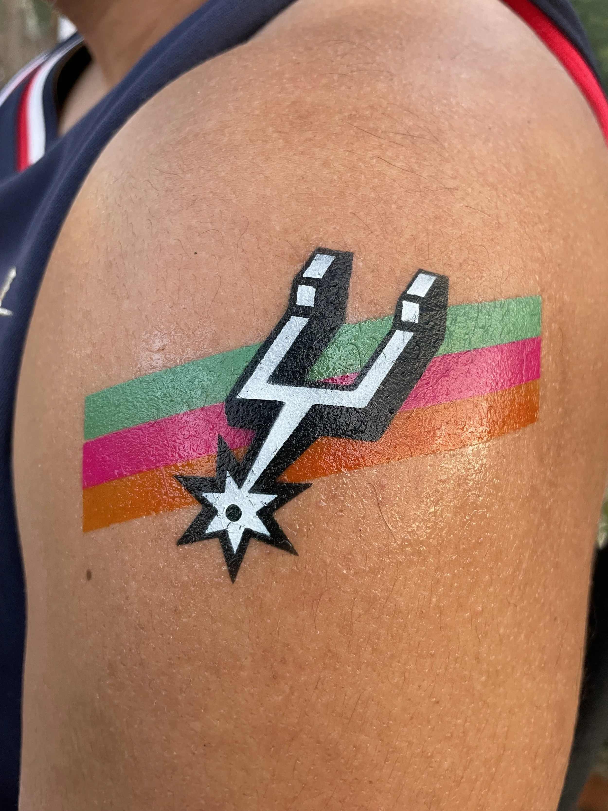 Tattoos on person's shoulder with a stylized lightning bolt with a star and colorful horizontal stripes in pink, orange, green, and black.