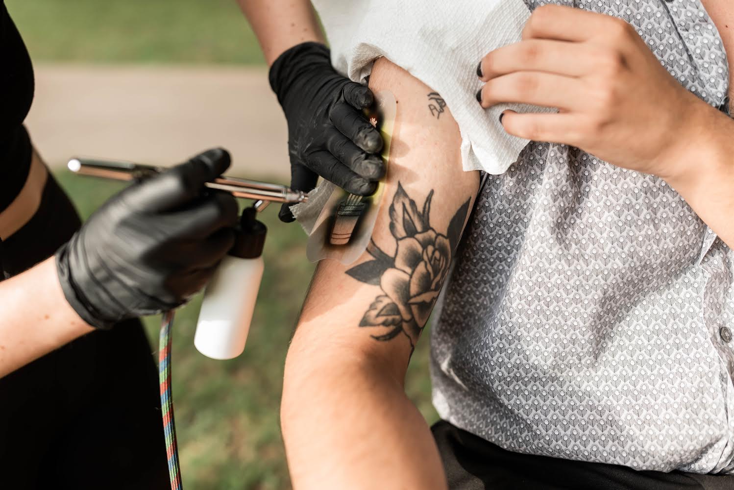 Person with tattoos on their arm getting an airbrush tattoo done by an artist wearing black gloves, outdoors.