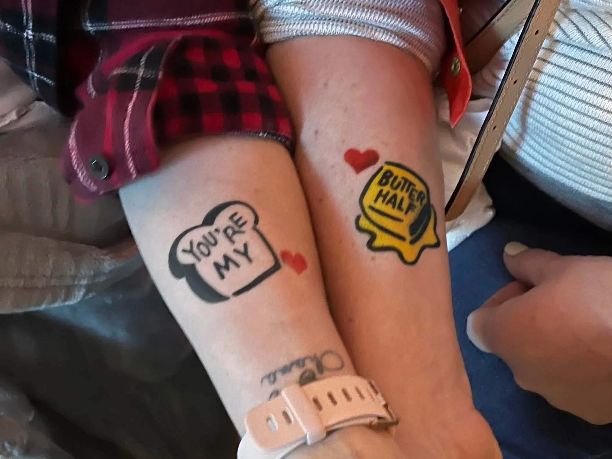 Close-up of a person's arm with visible tattoos, including a red heart, a yellow glove, and text, with a hospital wristband, and part of a plaid shirt sleeve.