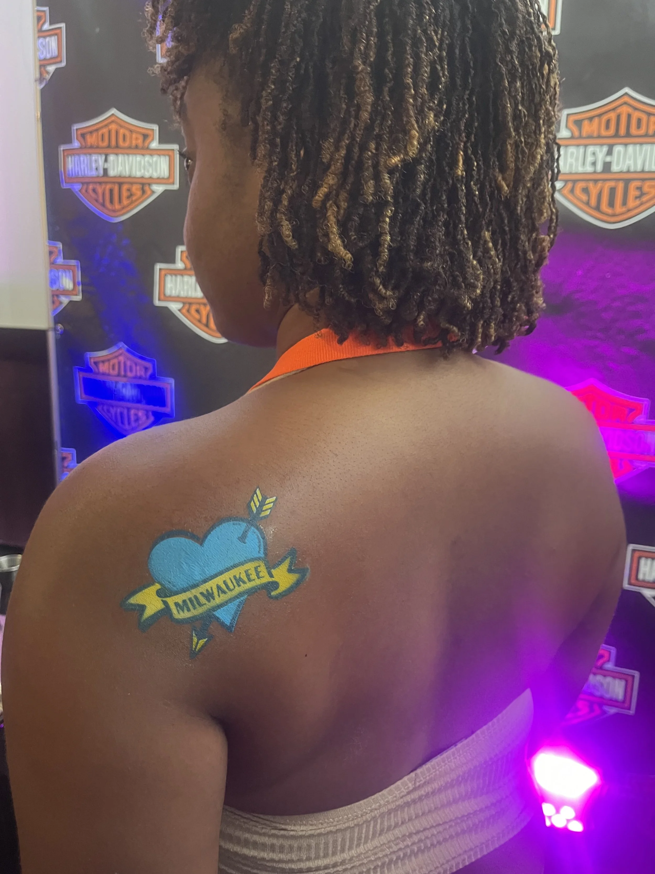 Back of a woman with dreadlocks and a tattoo of a blue heart with a yellow banner that says 'Milwaukee' on her shoulder, standing in front of a Harley-Davidson branded backdrop with purple and pink lighting.