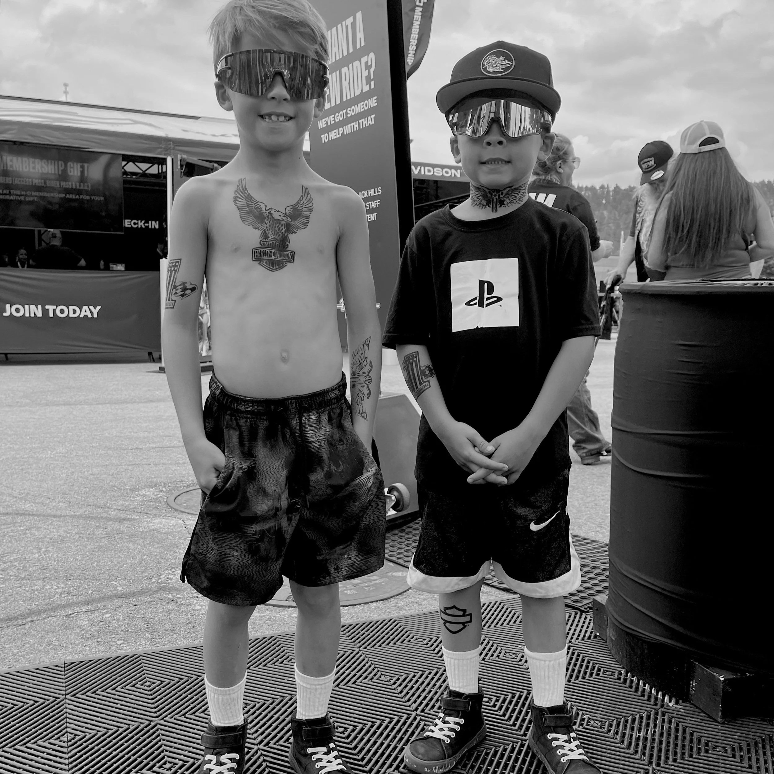 Two young boys standing side by side at an outdoor event, both wearing sunglasses and shorts. The boy on the left is shirtless, and the boy on the right is wearing a cap, a t-shirt with a PlayStation logo, and sneakers. There are other people and a tent in the background.