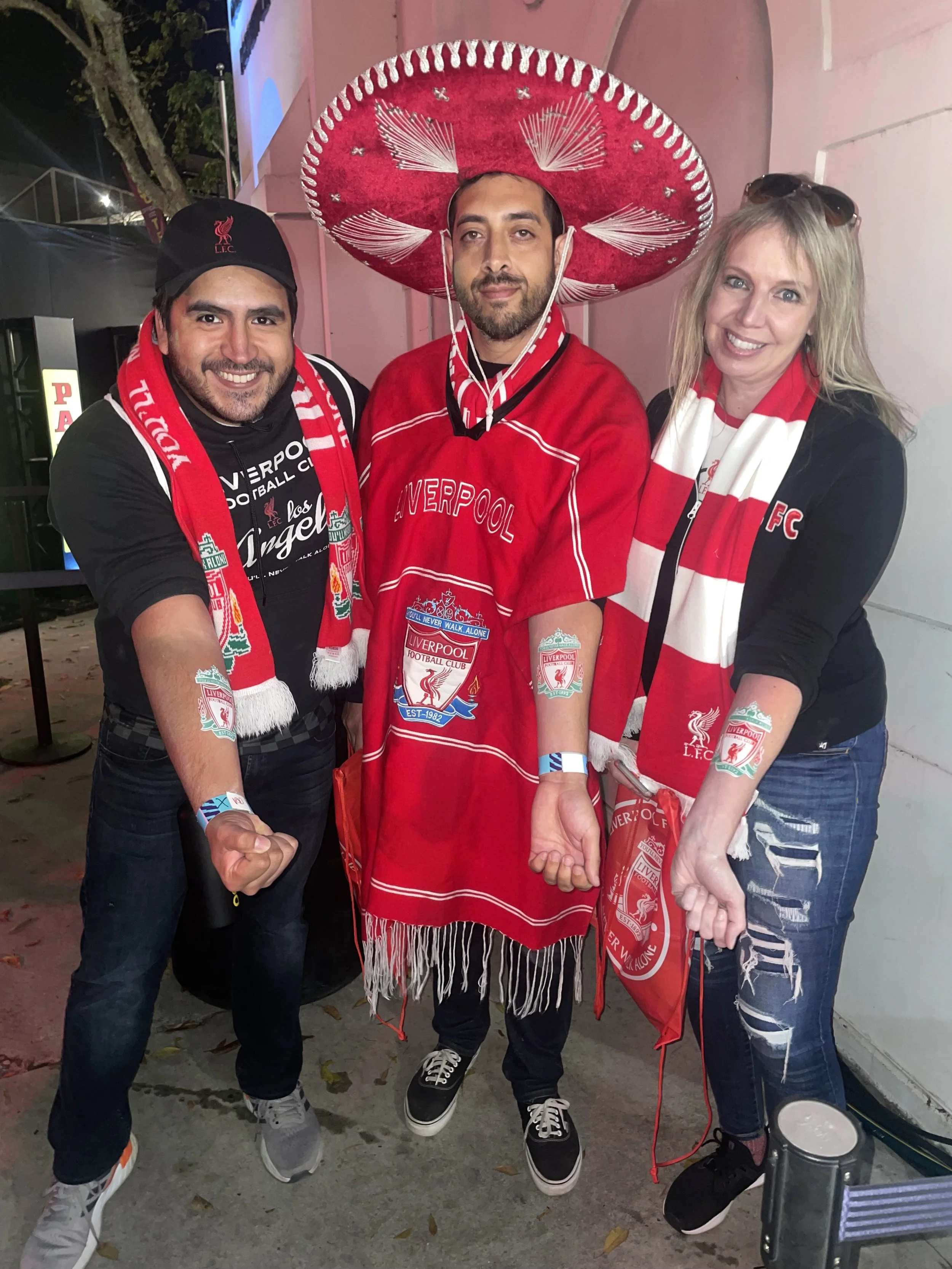 Three people celebrating Liverpool Football Club, two wearing scarves and one wearing a large red sombrero. They are holding hands in a show of unity and support.