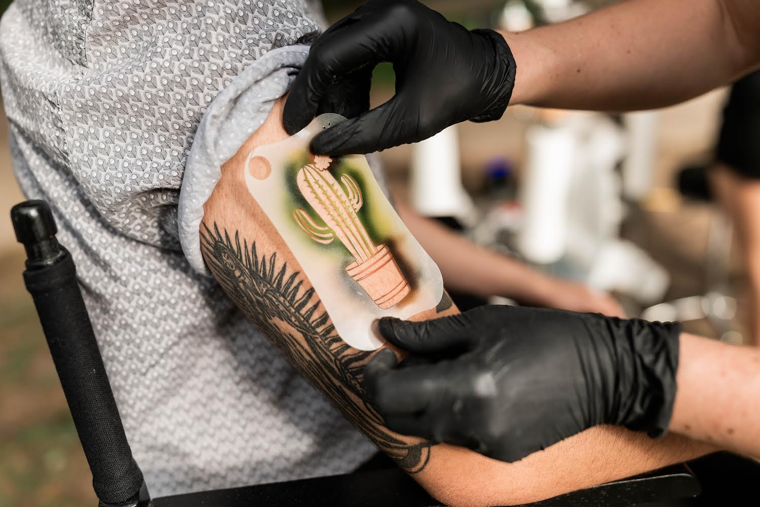 A tattoo artist is applying a cactus-themed tattoo to a person's forearm. The artist is wearing black gloves and holding a stencil for the tattoo.