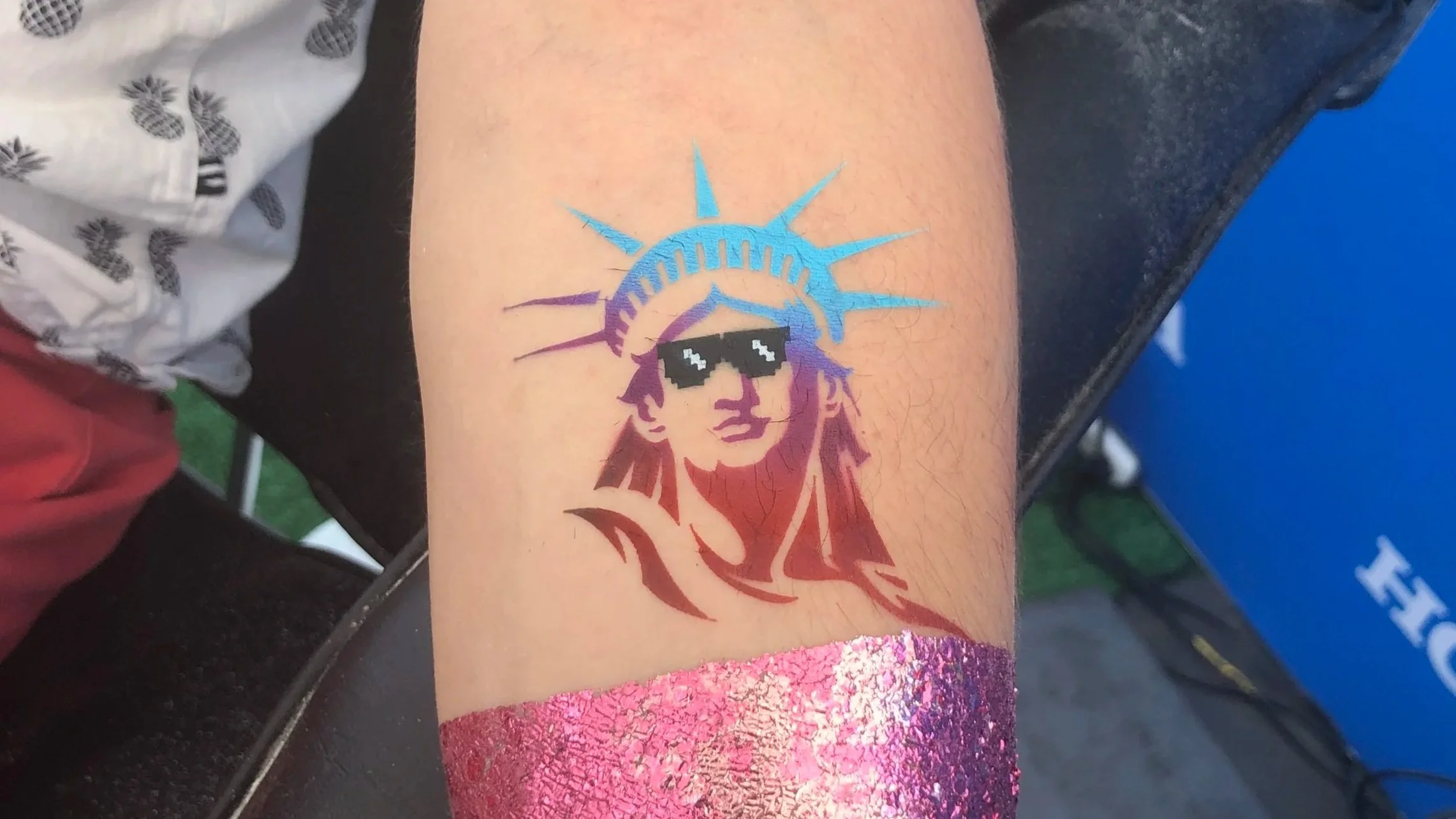 A tattoo of the Statue of Liberty with sunglasses on a person's arm, with pink glitter nail polish visible at the bottom.