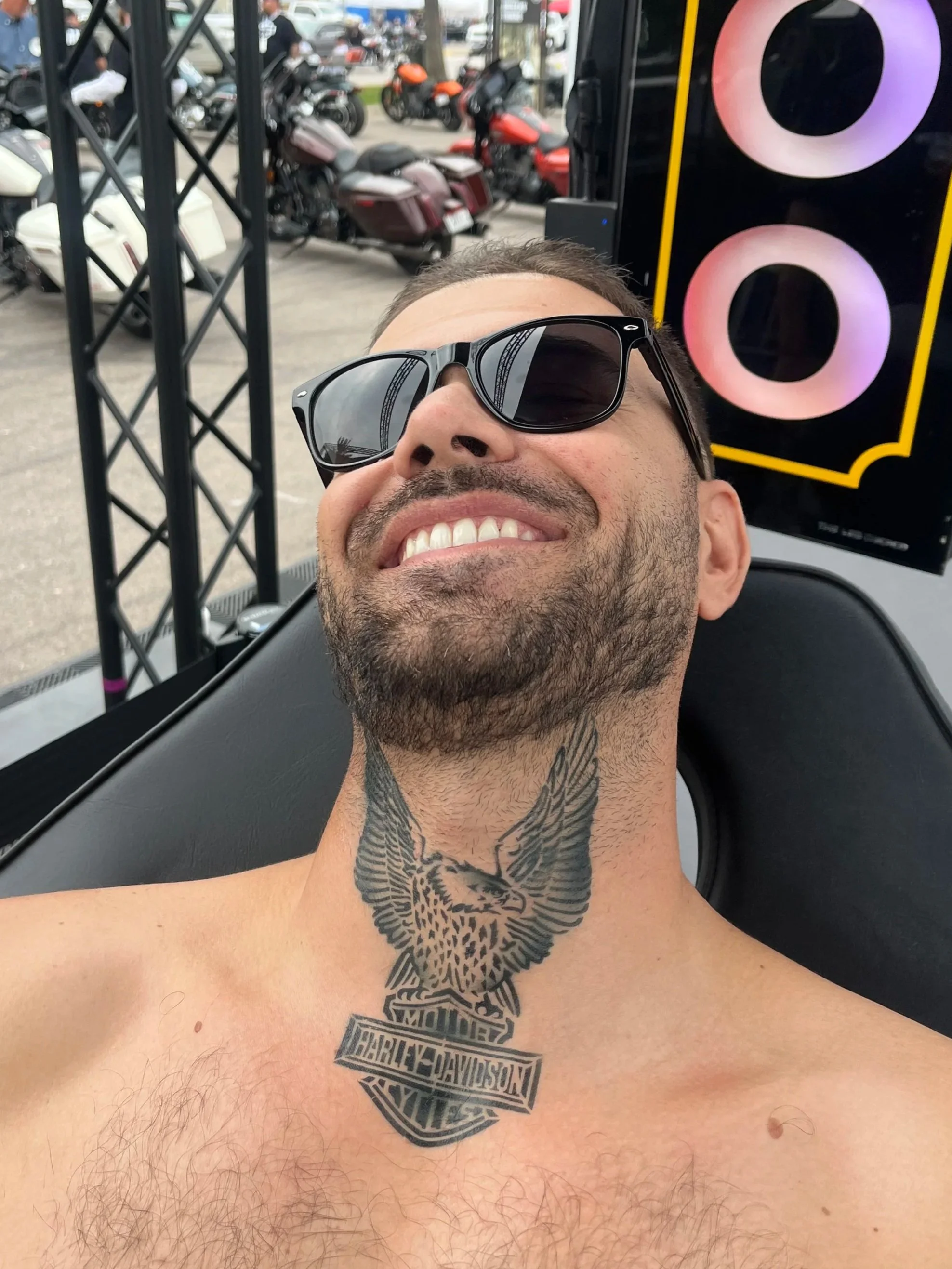 A man with sunglasses taking a selfie, showing his tattooed chest with a Harley-Davidson logo tattooed across it and a detailed owl tattooed on his neck, in front of a display with motorcycles in the background.
