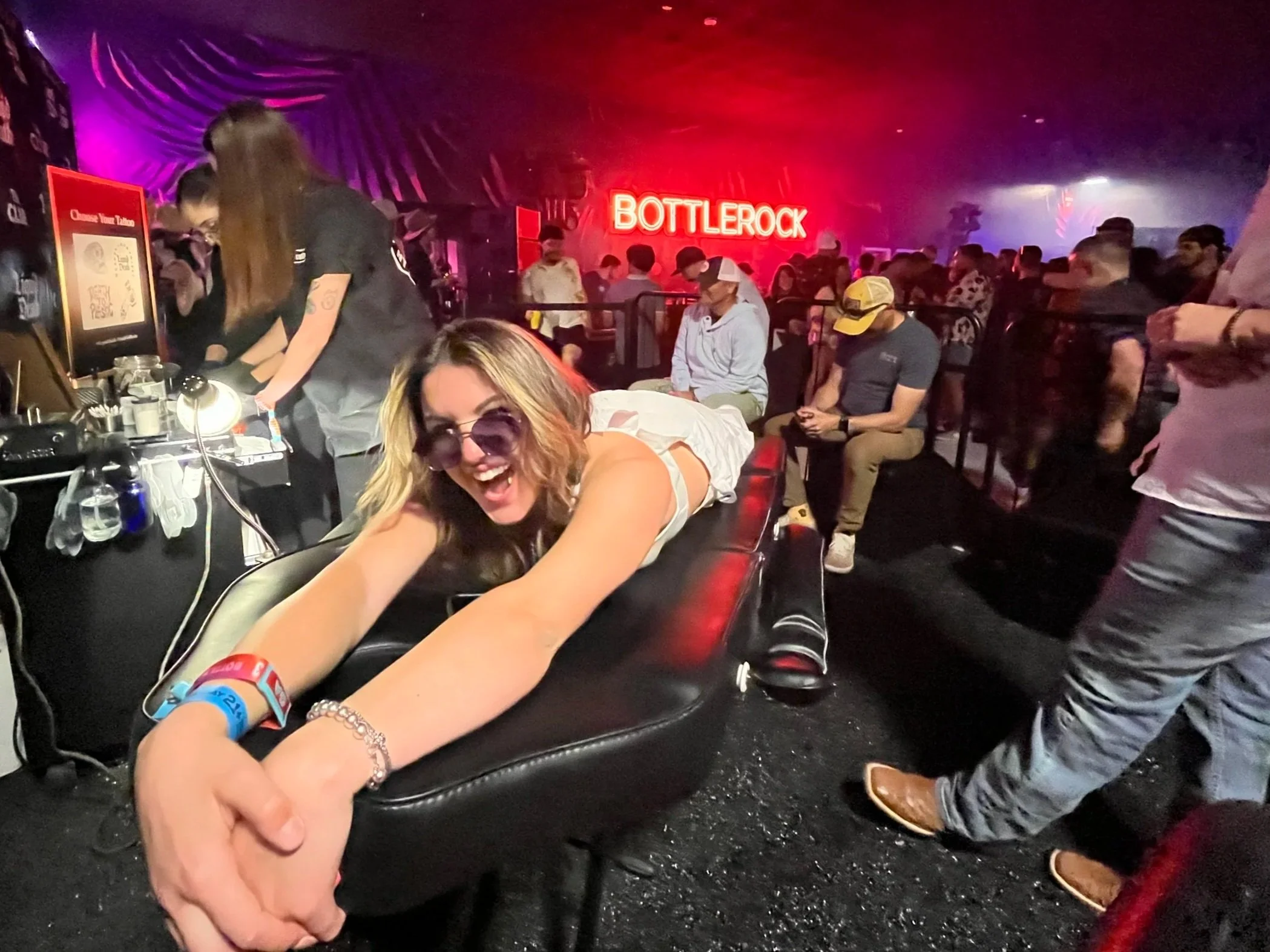 Woman smiling and leaning on a table with people sitting in a nightclub or concert venue with a red neon "Bottlerock" sign in the background.