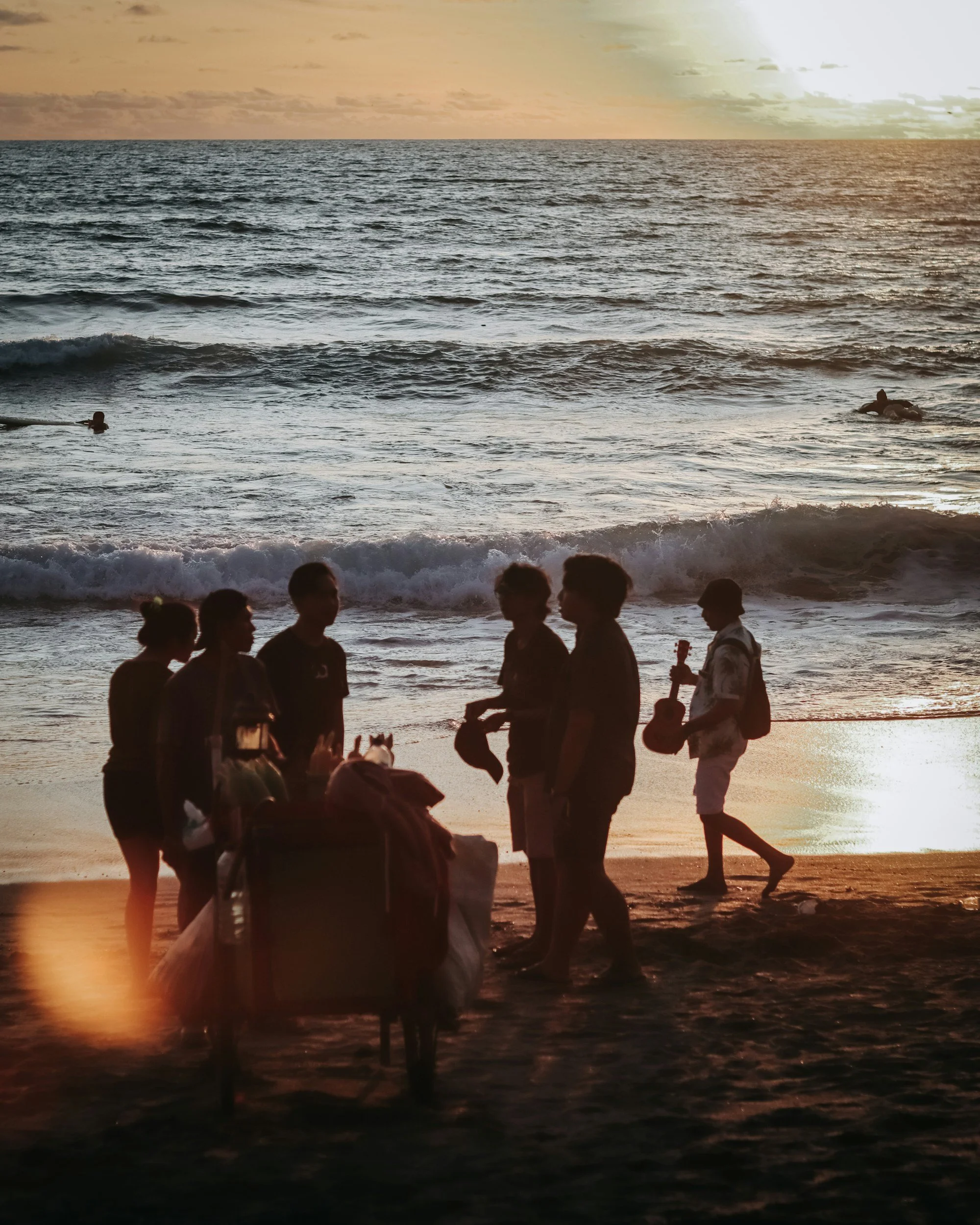 Silhouettes of people on a beach during sunset, some holding surfboards, with the ocean and a cloudy sky in the background.
