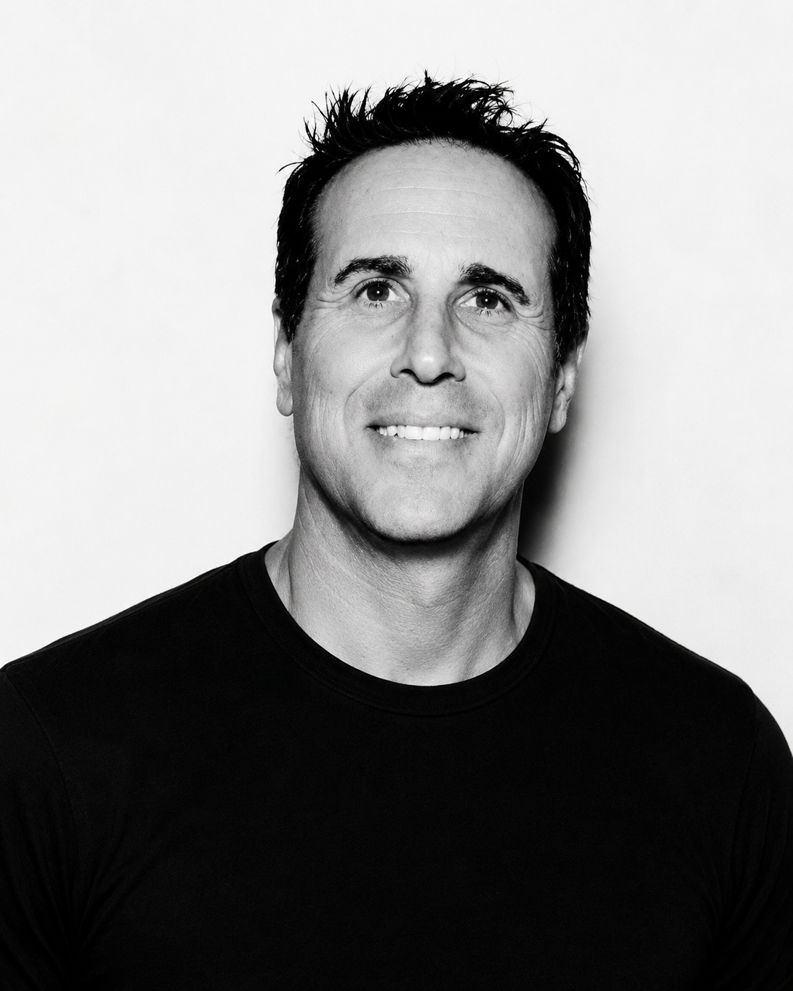 Black and white portrait of a smiling man with short dark hair, wearing a black shirt, looking directly at the camera against a plain white background.