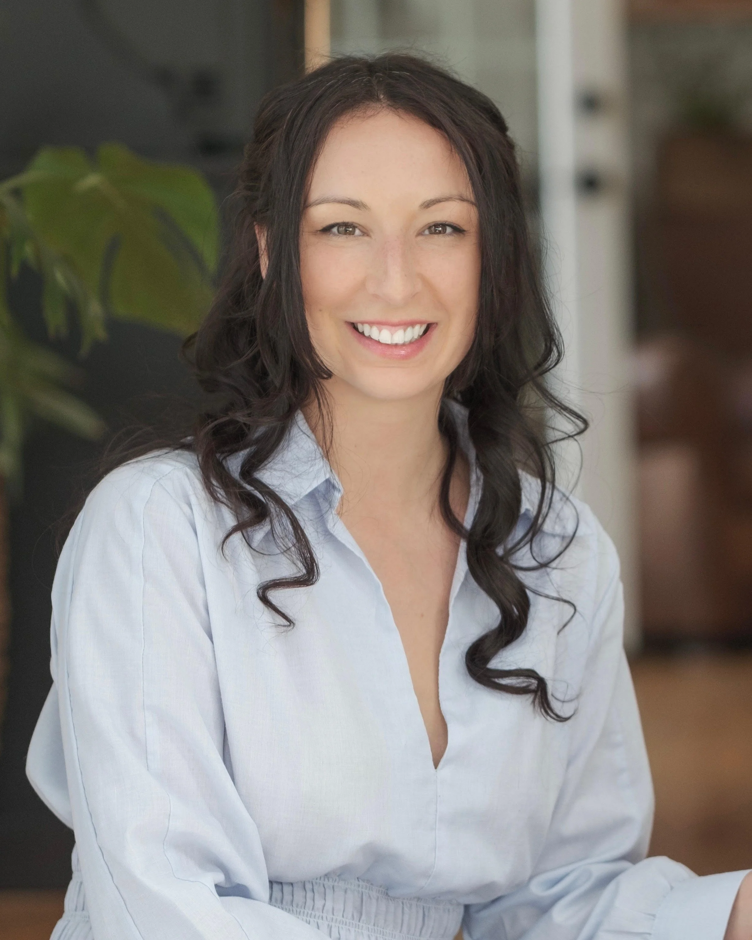 A woman with dark wavy hair smiling and wearing a light blue button-up shirt.
