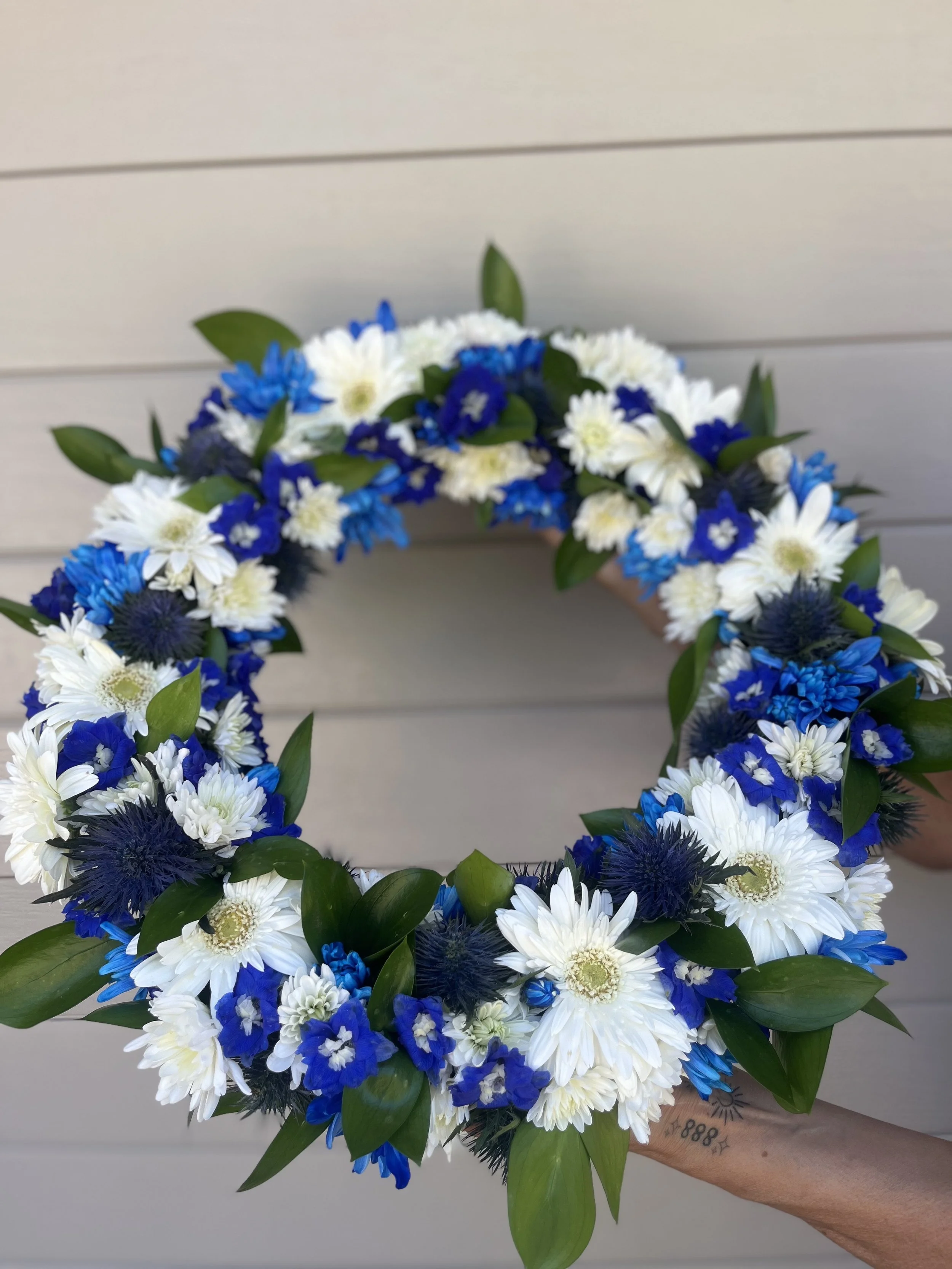 A floral wreath with white, blue, and dark purple flowers and green leaves, held by a person with a tattooed arm, against a light horizontal siding background.