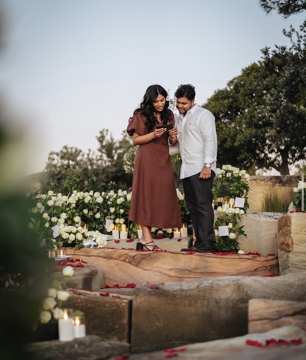 A young couple stands on a stone platform surrounded by white roses, candles, and rose petals, outdoors during sunset, looking at a phone and smiling.