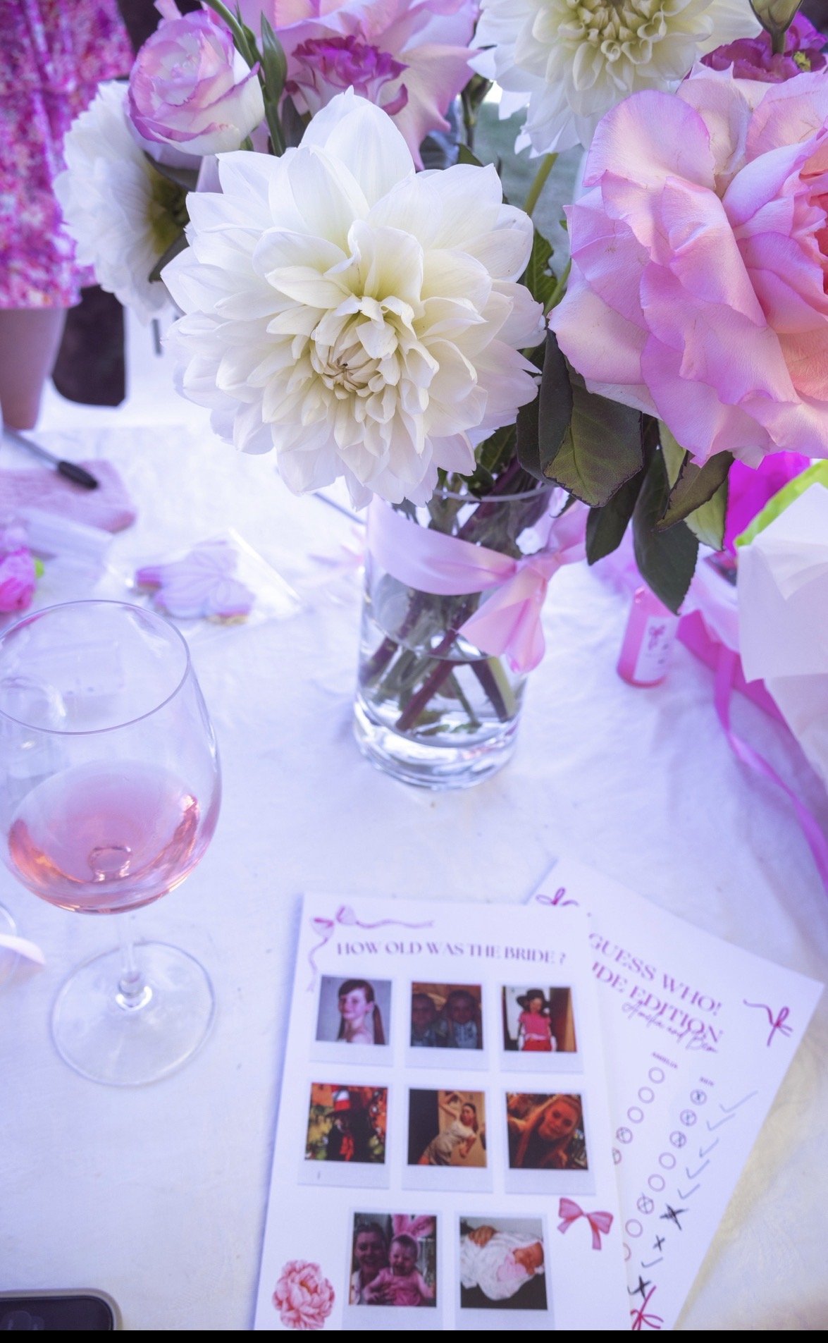 A bouquet of white, pink, and cream-colored flowers in a glass vase on a table with a pink ribbon tied around the vase. There is a glass of pink beverage and a piece of paper with photo booth pictures and pink decorations on the table.
