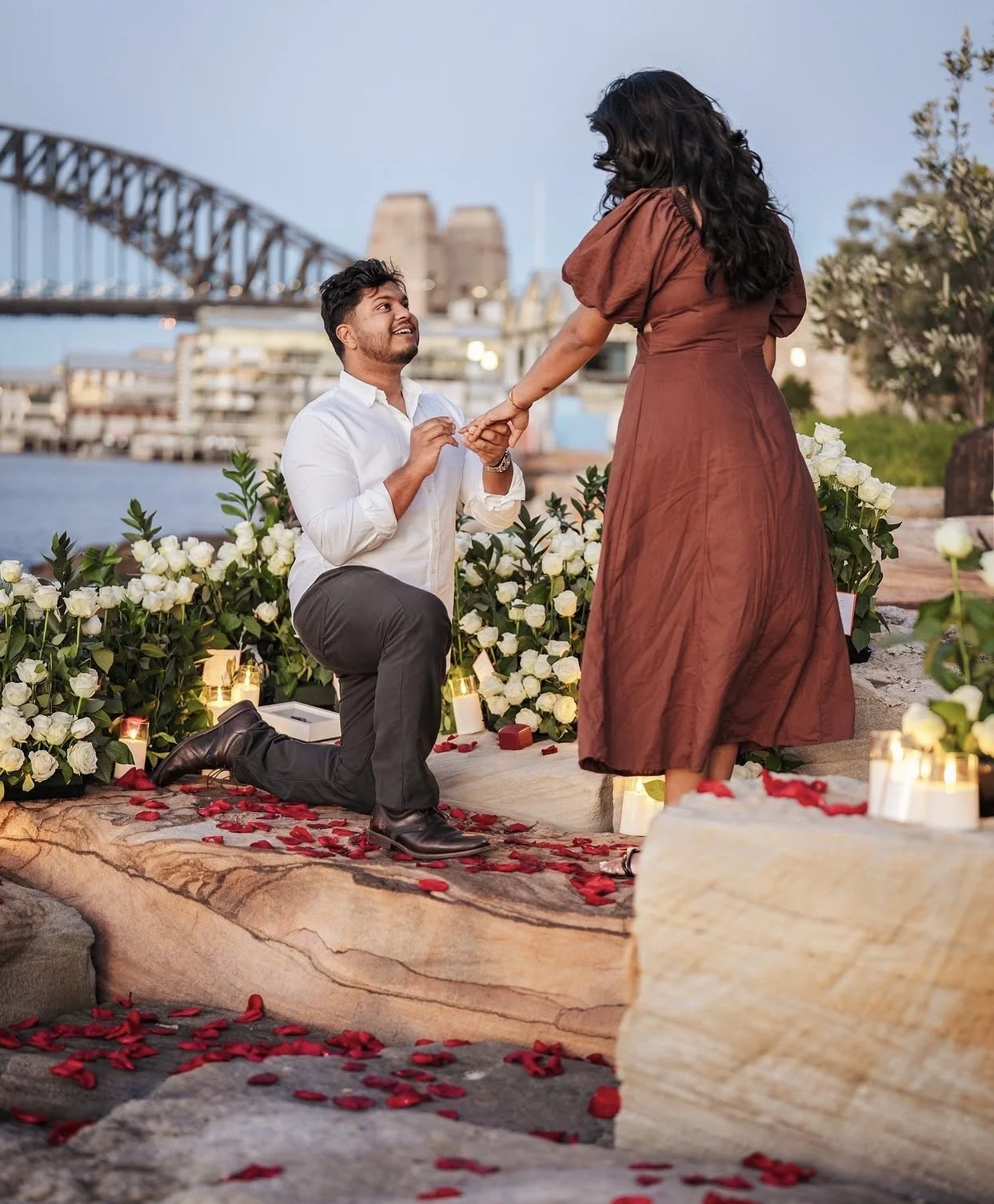 A man proposes marriage to a woman on a romantic outdoor setup near a river, with the Sydney Harbour Bridge in the background. The scene includes rose petals, candles, and white flowers.