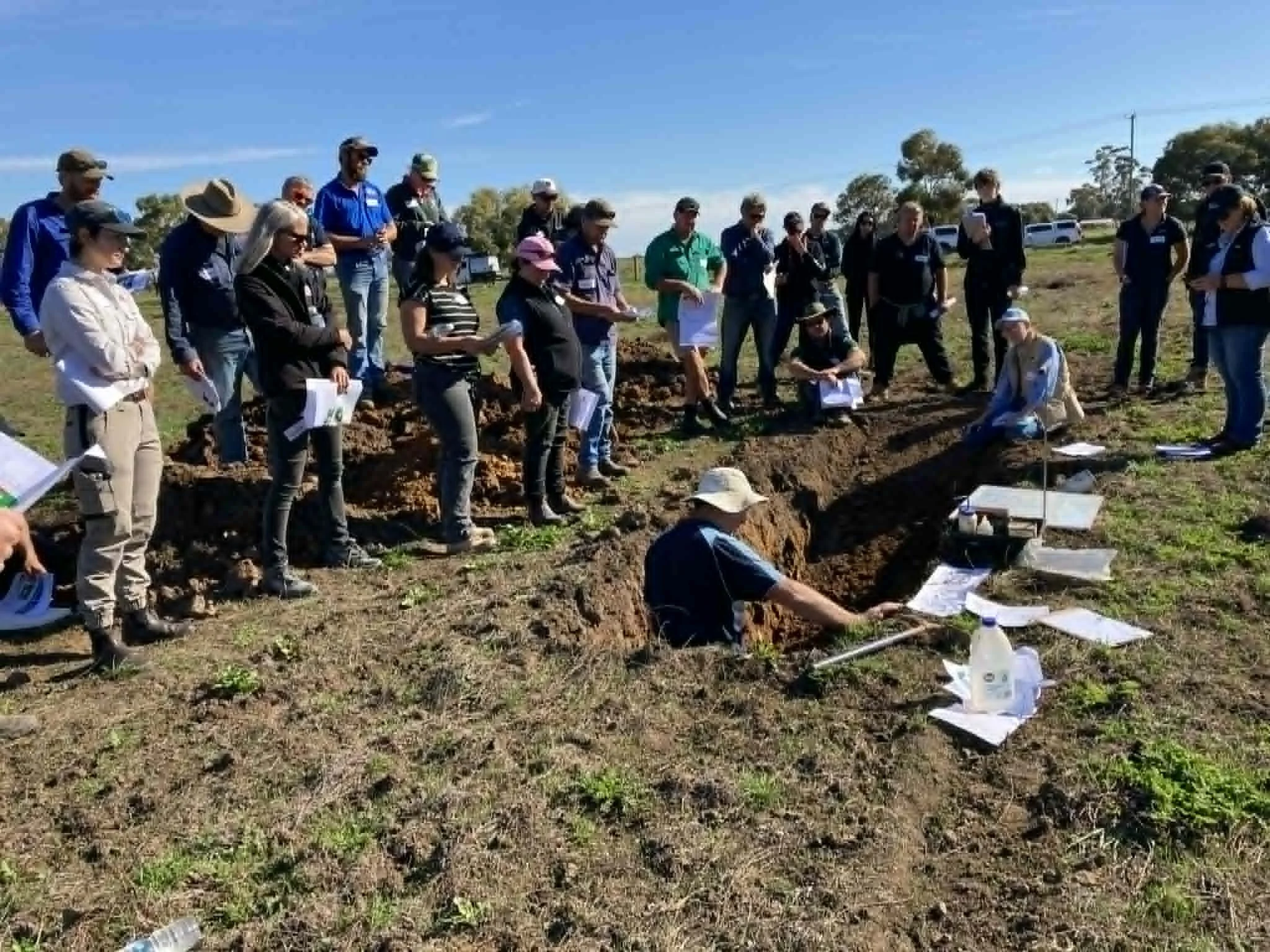 A group of people standing outdoors on a sunny day, gathered around a trench or excavation site during a field event or presentation.