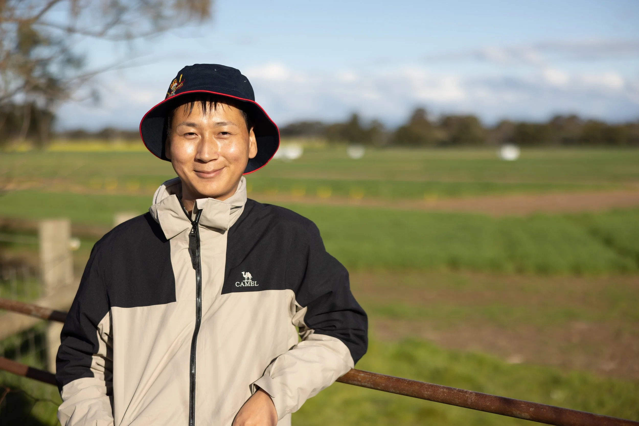 A man smiling outdoors wearing a black and beige jacket, standing near a wooden fence on a green field with trees and a cloudy sky in the background.