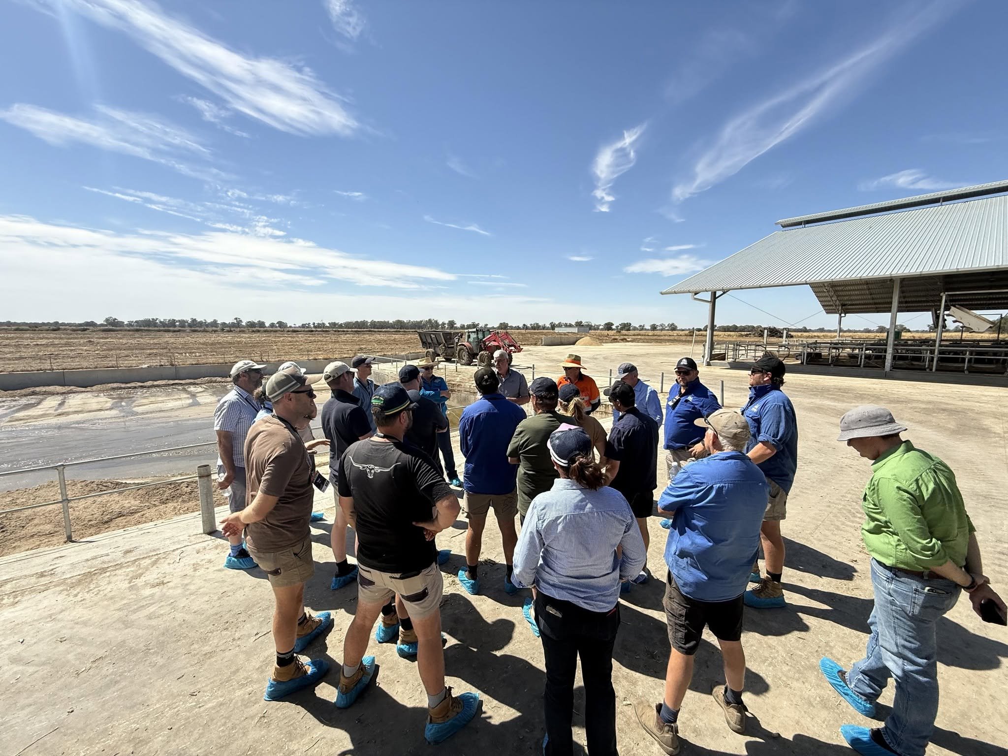 A group of people wearing blue shoe covers gathered outdoors near a large metal-roofed structure and a tractor, with a clear blue sky above and open land in the background.