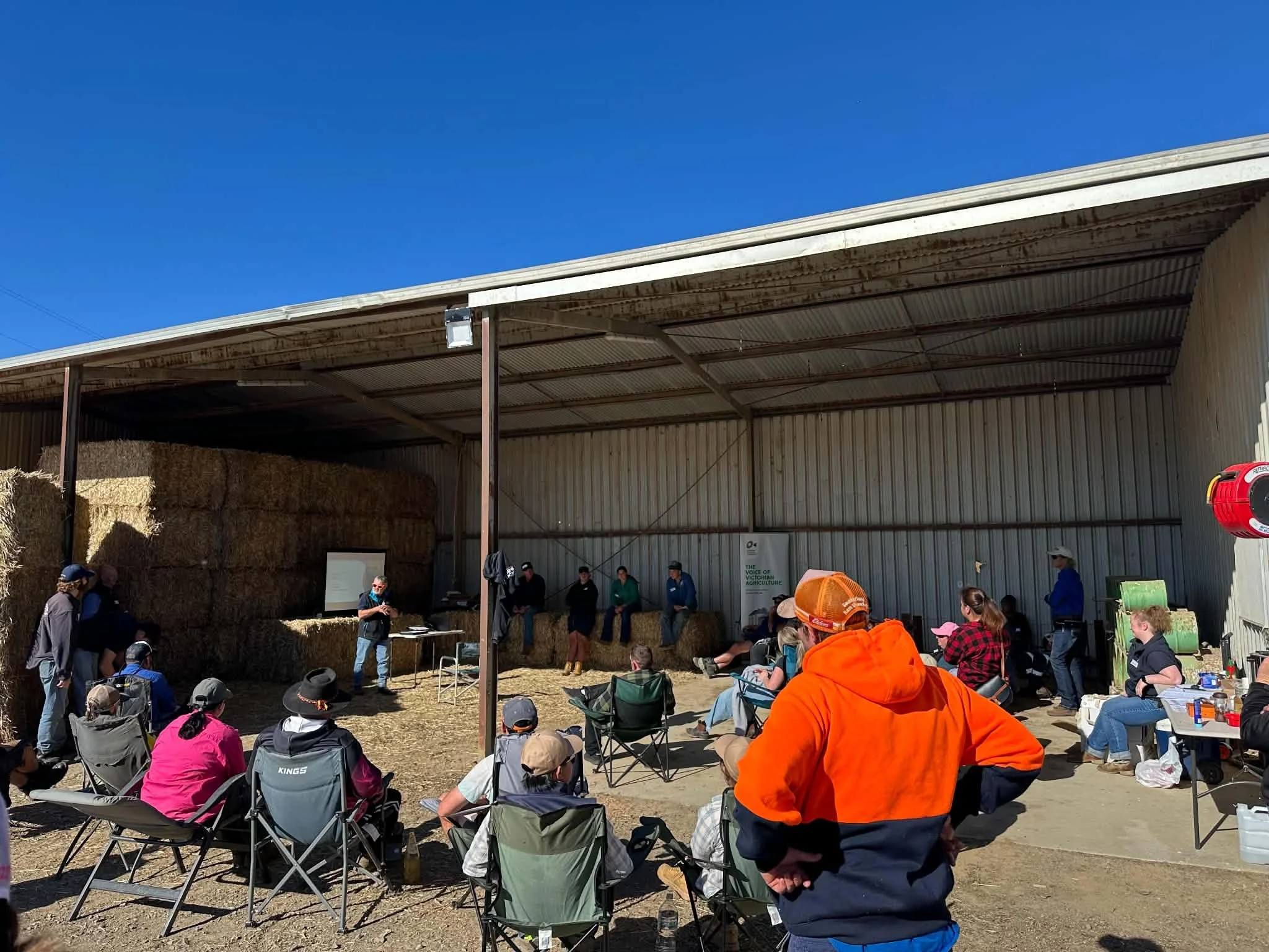 People gathered outdoors in front of a large shed with hay bales inside, attending a presentation or demonstration under a sunny, clear blue sky.