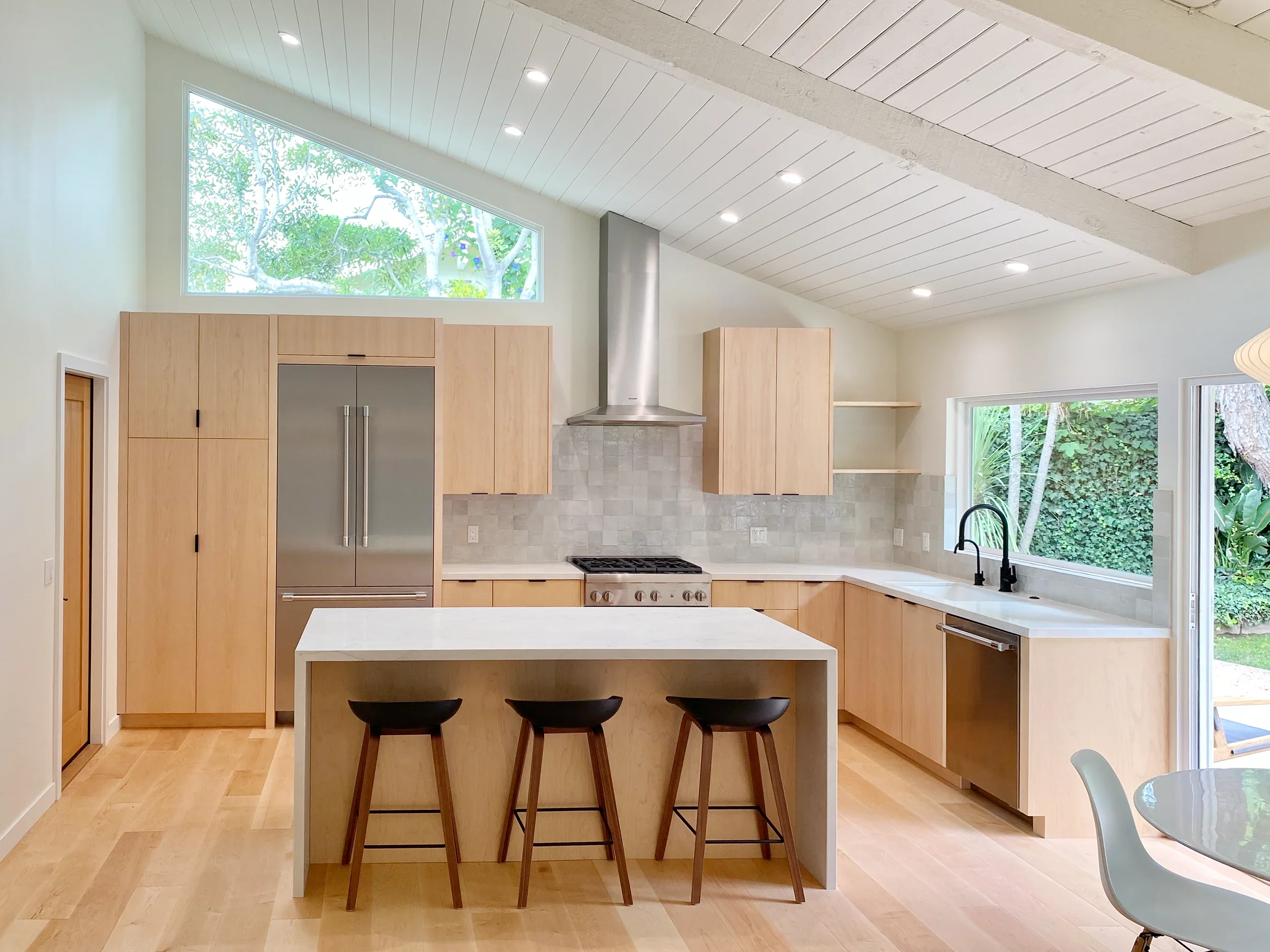 Modern kitchen with light wood cabinets, a white island, a stainless steel refrigerator, a stove with a stainless steel hood, large windows showing outdoor greenery, a high sloped ceiling with recessed lighting, and a small round dining table with a gray chair.
