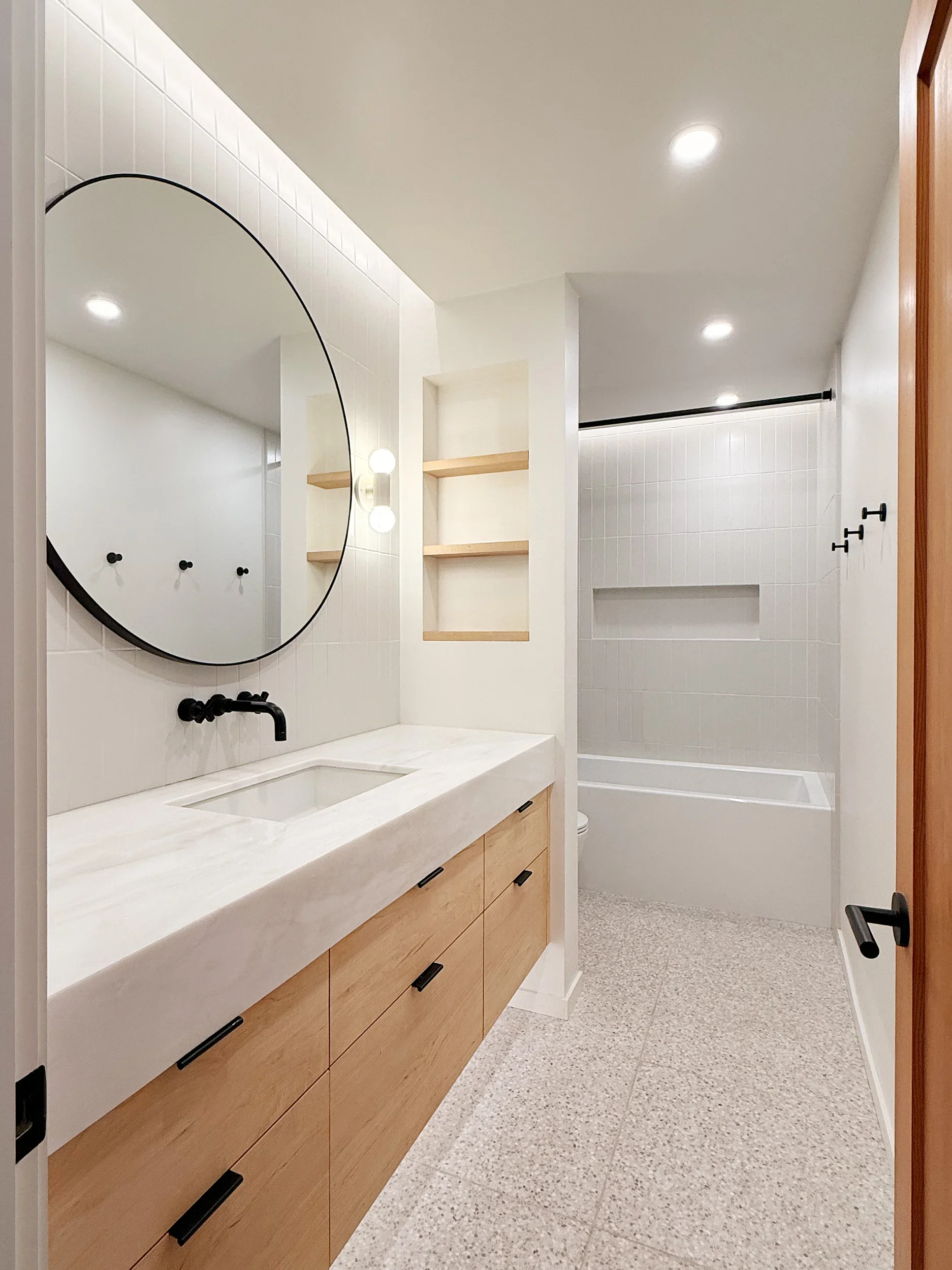 A modern bathroom with a marble countertop, sink, black faucet, maple wood cabinets, a large round mirror, open wooden shelves, white tiled shower area with a black shower rod, and a speckled beige floor.