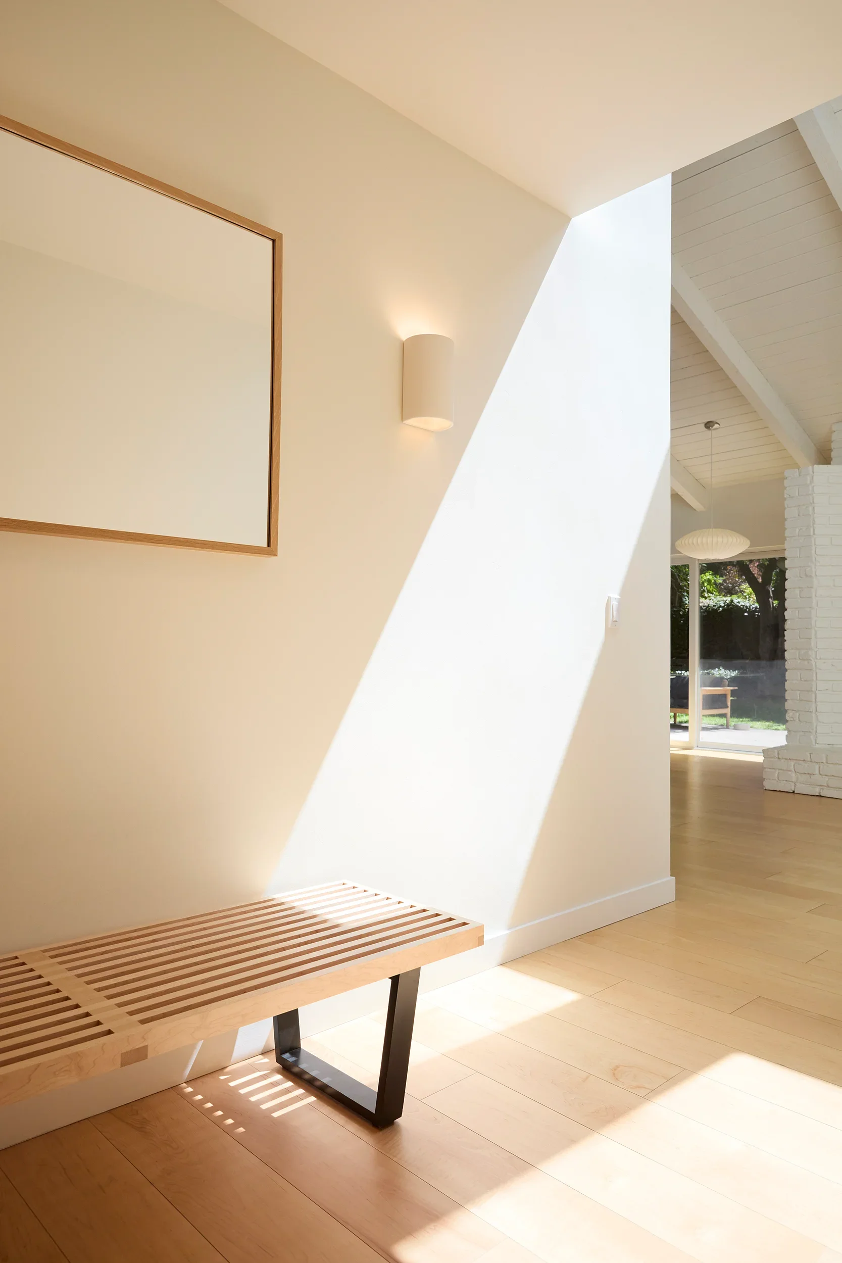 Bright interior space with sunlight casting shadows on light wooden floor, white walls, a beige high vaulted ceiling, and a small hallway with a hanging light fixture and large window showing greenery outside.