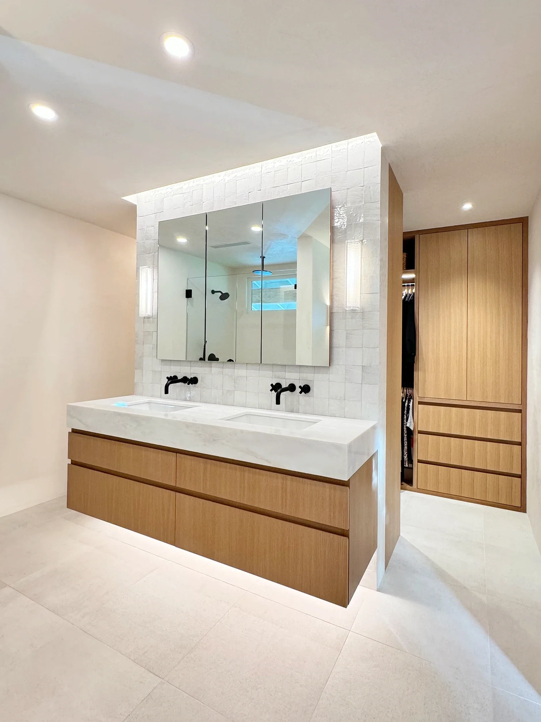 Modern bathroom with double vanity with white oak wood cabinets, large mirror, black faucet, and white textured wall, with built-in wooden storage on the right side.