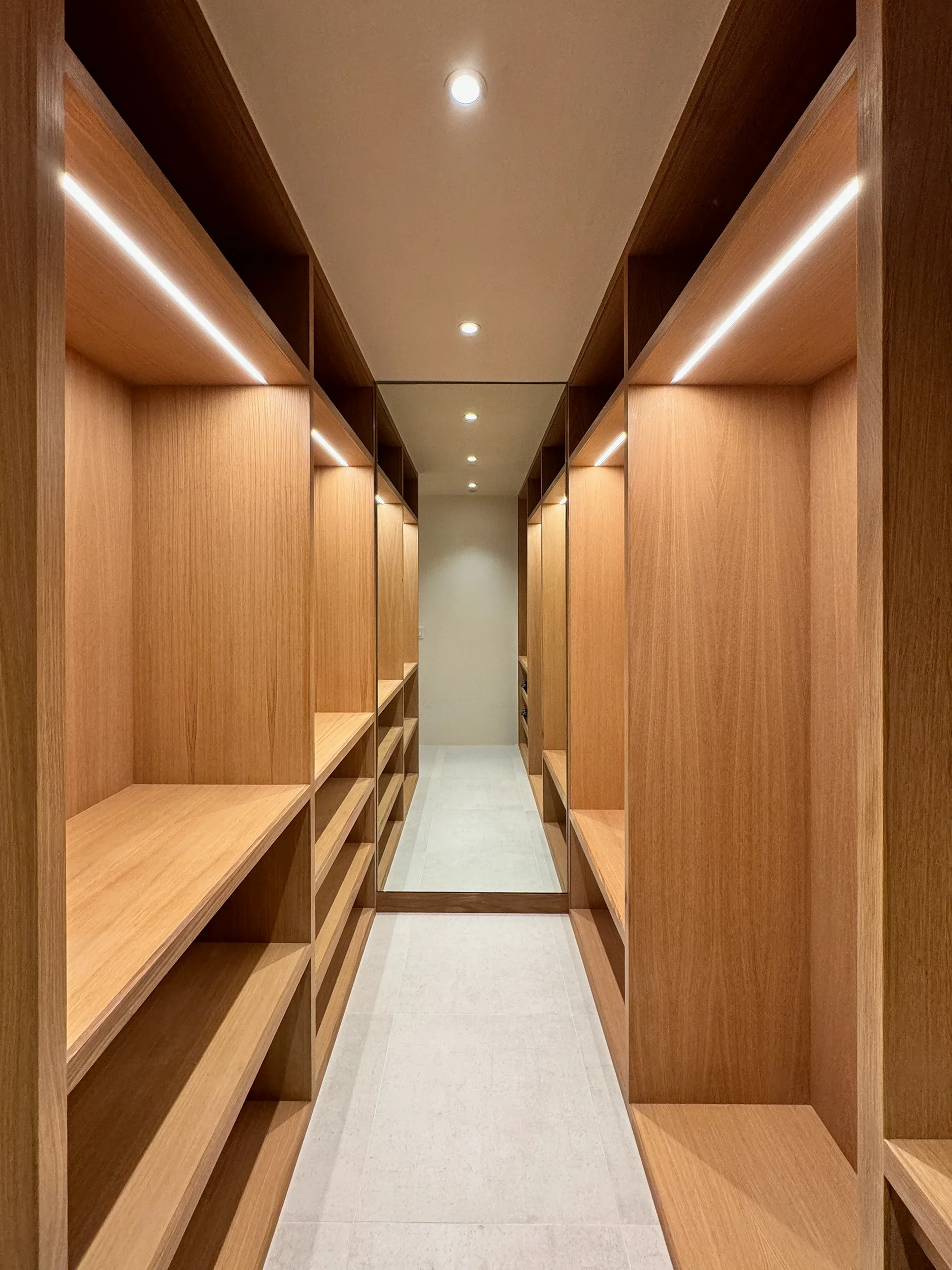 White Oak wooden walk-in closet with built-in shelves and a large mirror at the end, illuminated by ceiling lights.