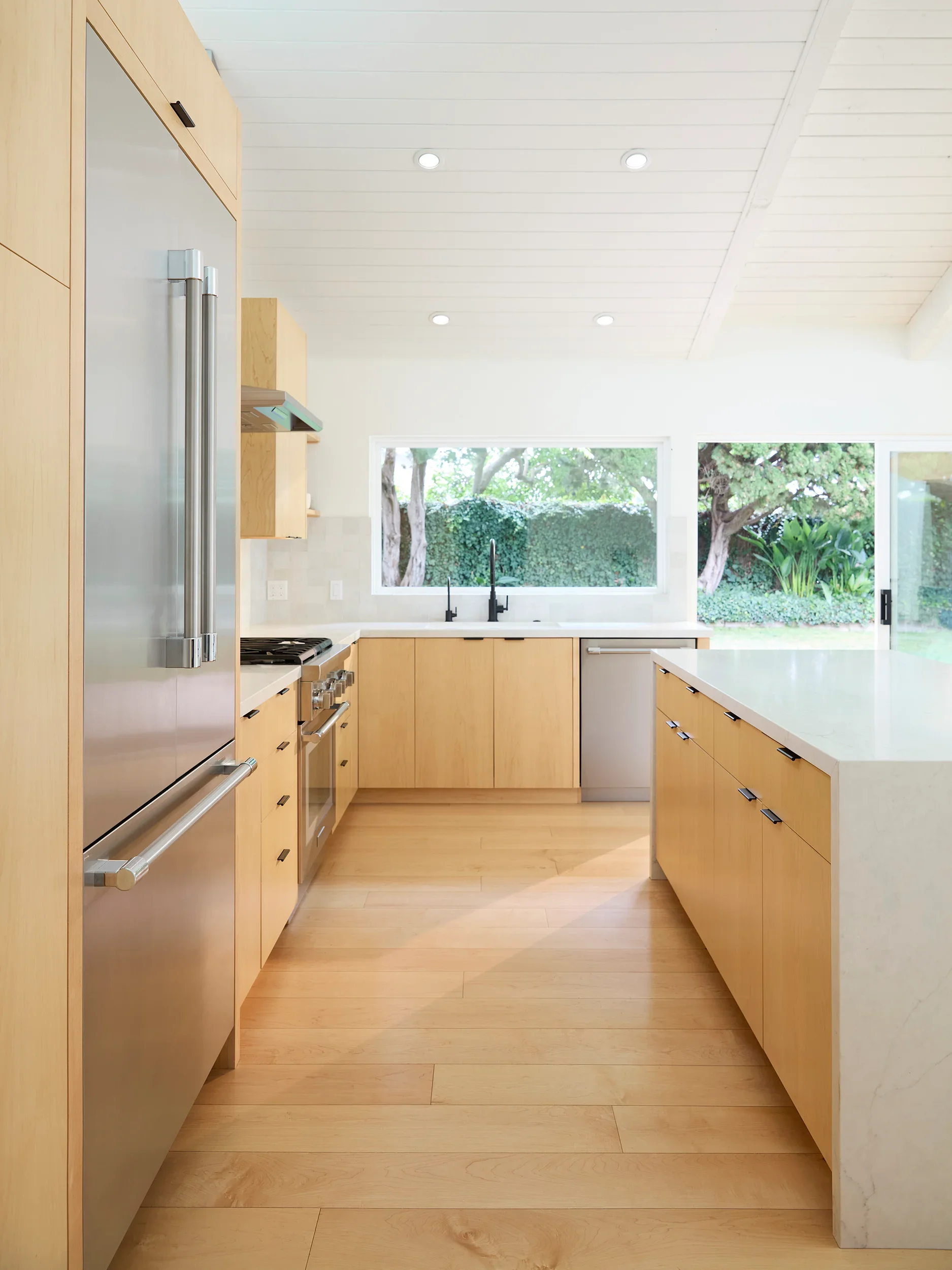 Modern kitchen with maple wood cabinets, white countertops, stainless steel appliances, large window overlooking greenery, and light wood flooring.
