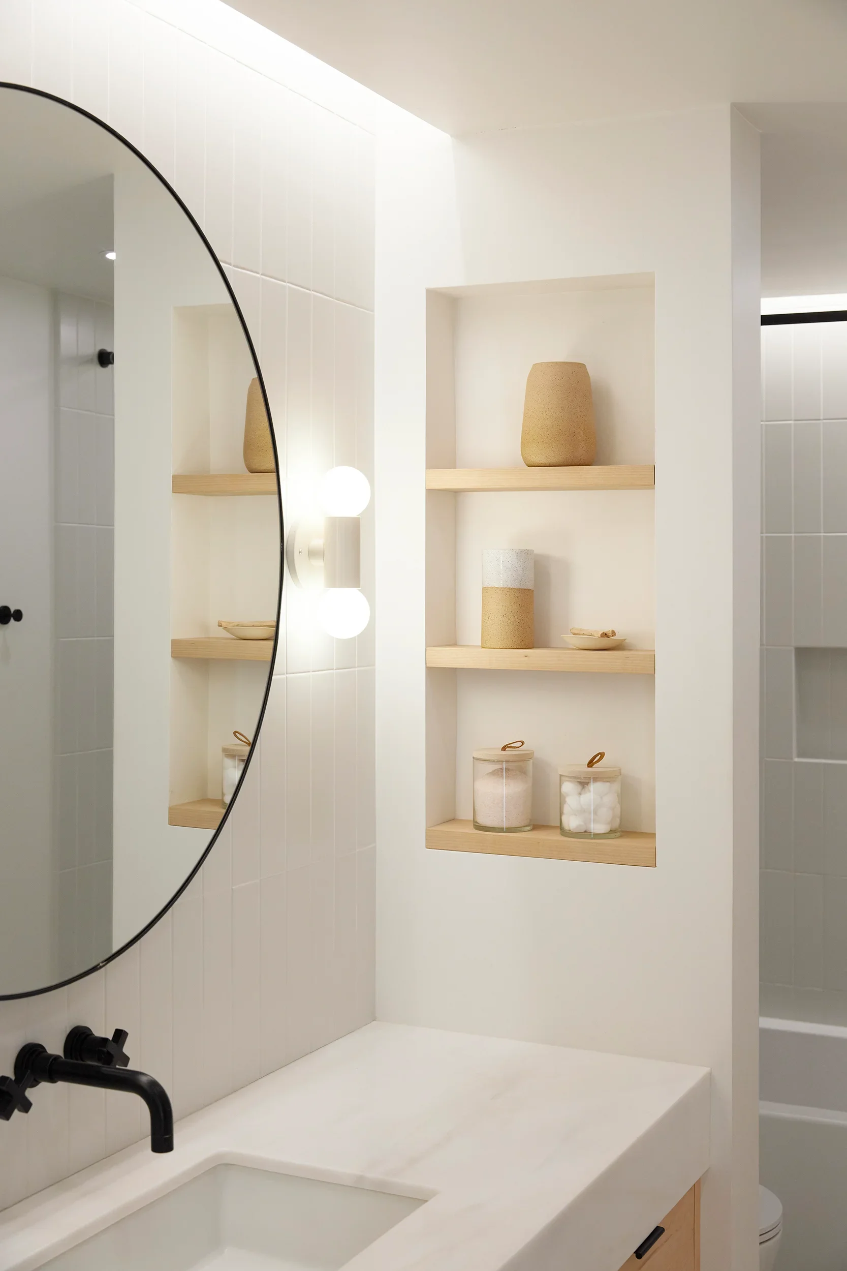 A modern bathroom sink area with a wmarble countertop, a black faucet, a round mirror, and wooden shelves holding beige vases and small decorative items.