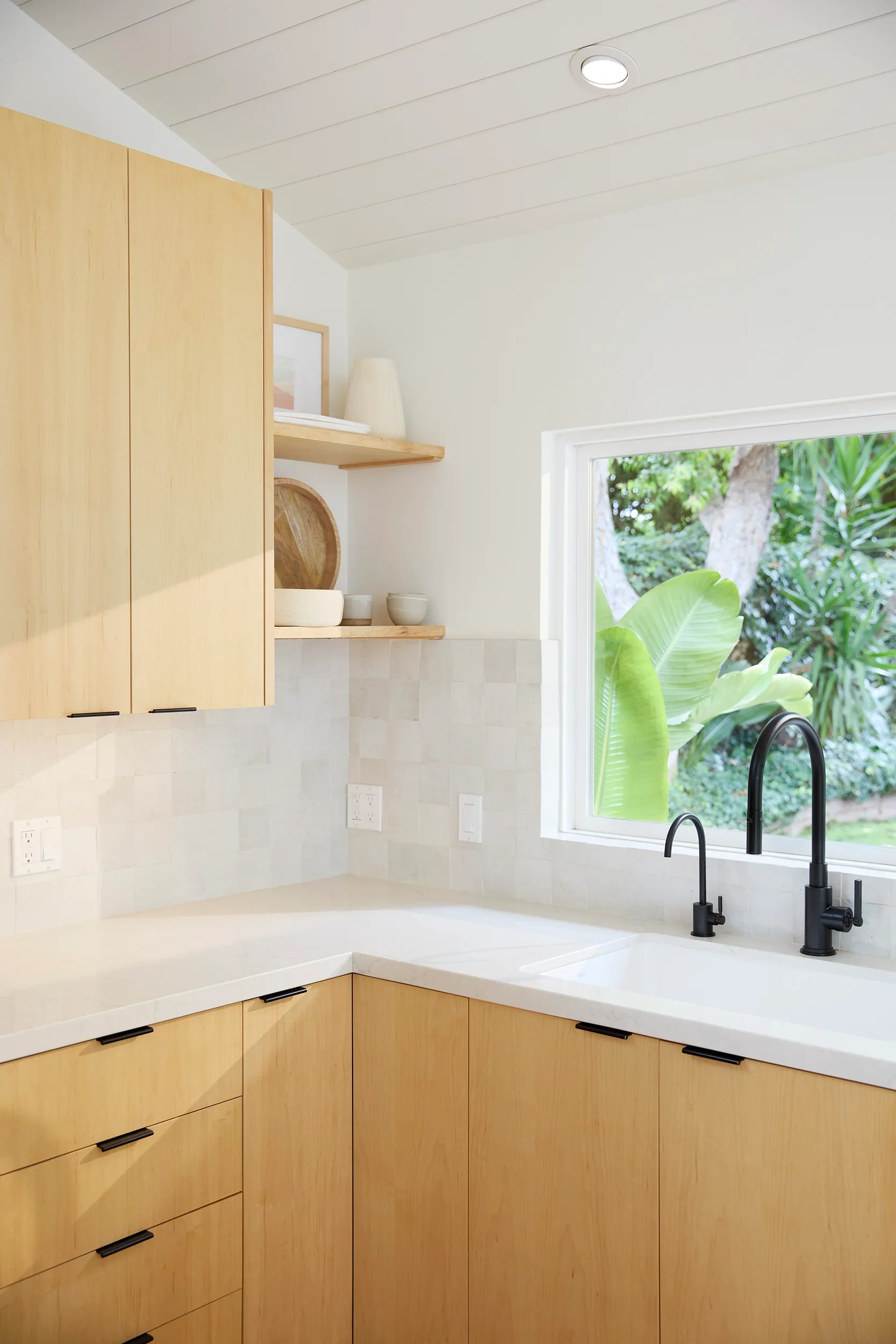 Kitchen corner with maple wood cabinets, open shelf with bowls and decor, window with a view of trees and plants outside, black faucet at kitchen sink, white countertop.
