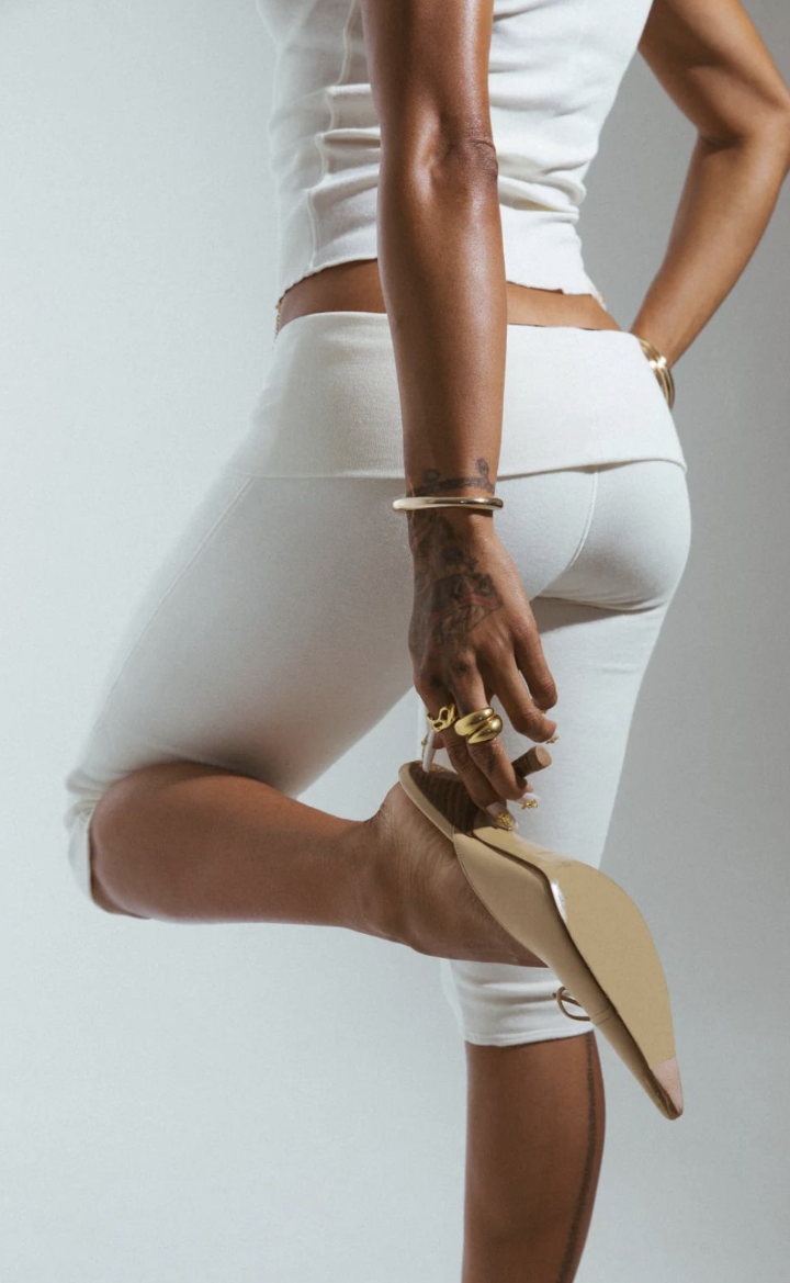 A woman wearing white pants and a sleeveless top, holding her foot with her right hand, showing gold jewelry and beige high-heeled shoes.